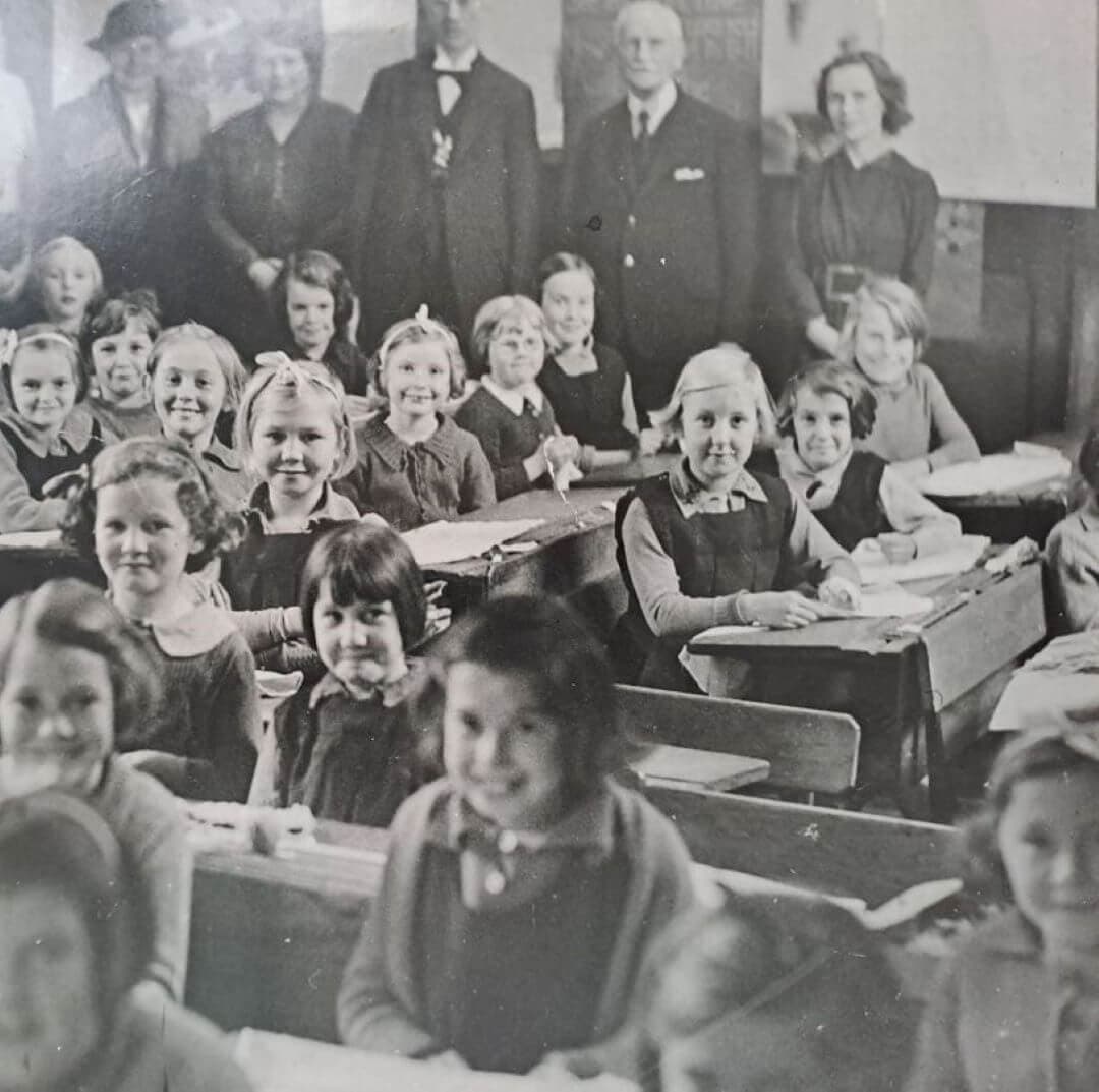 Vintage classroom with smiling children sitting at desks while teachers and adults stand at the back, observing. - Home Instead