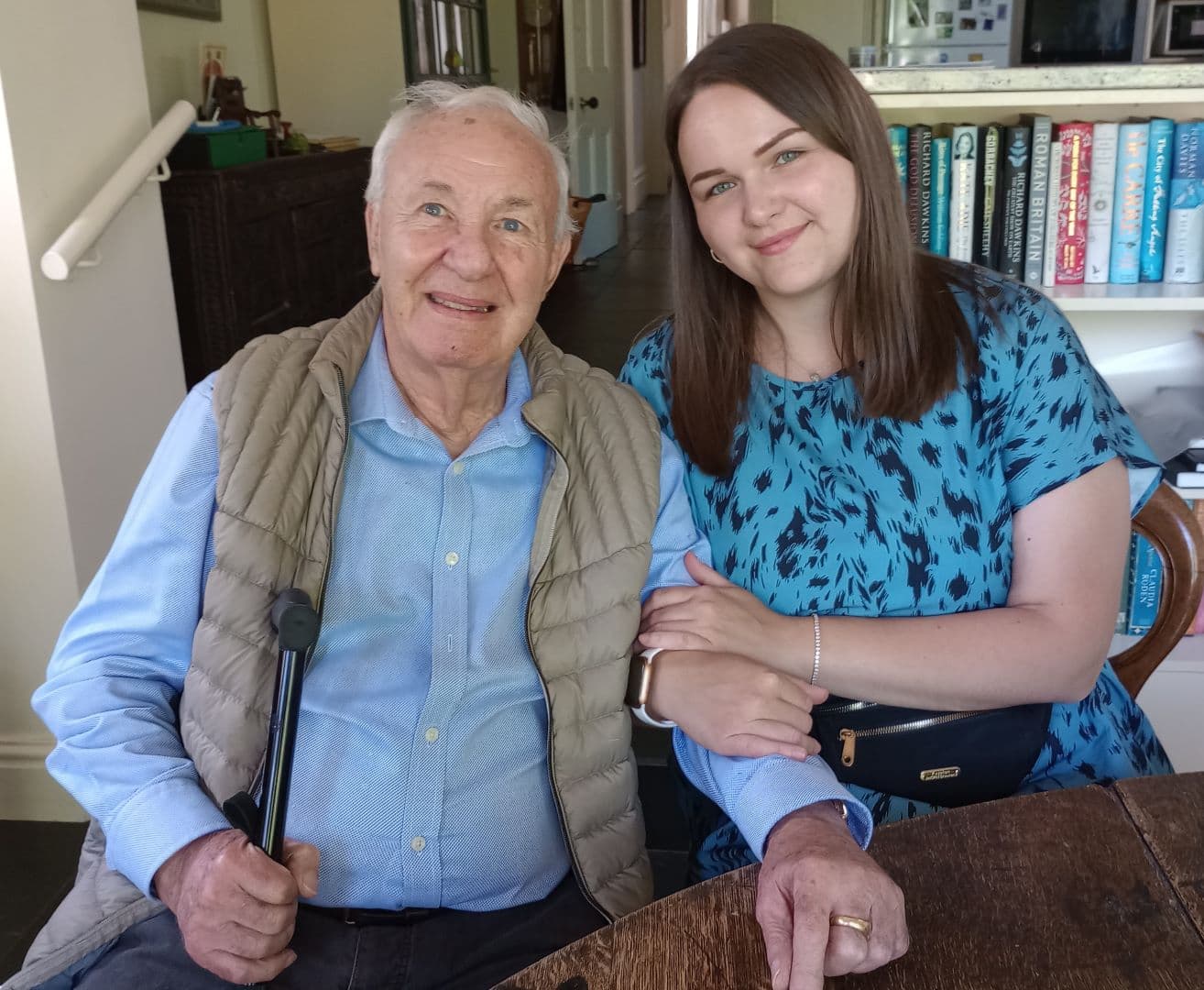 Elderly man sitting and holding a cane, with a young woman in a blue dress smiling and holding his arm. - Home Instead
