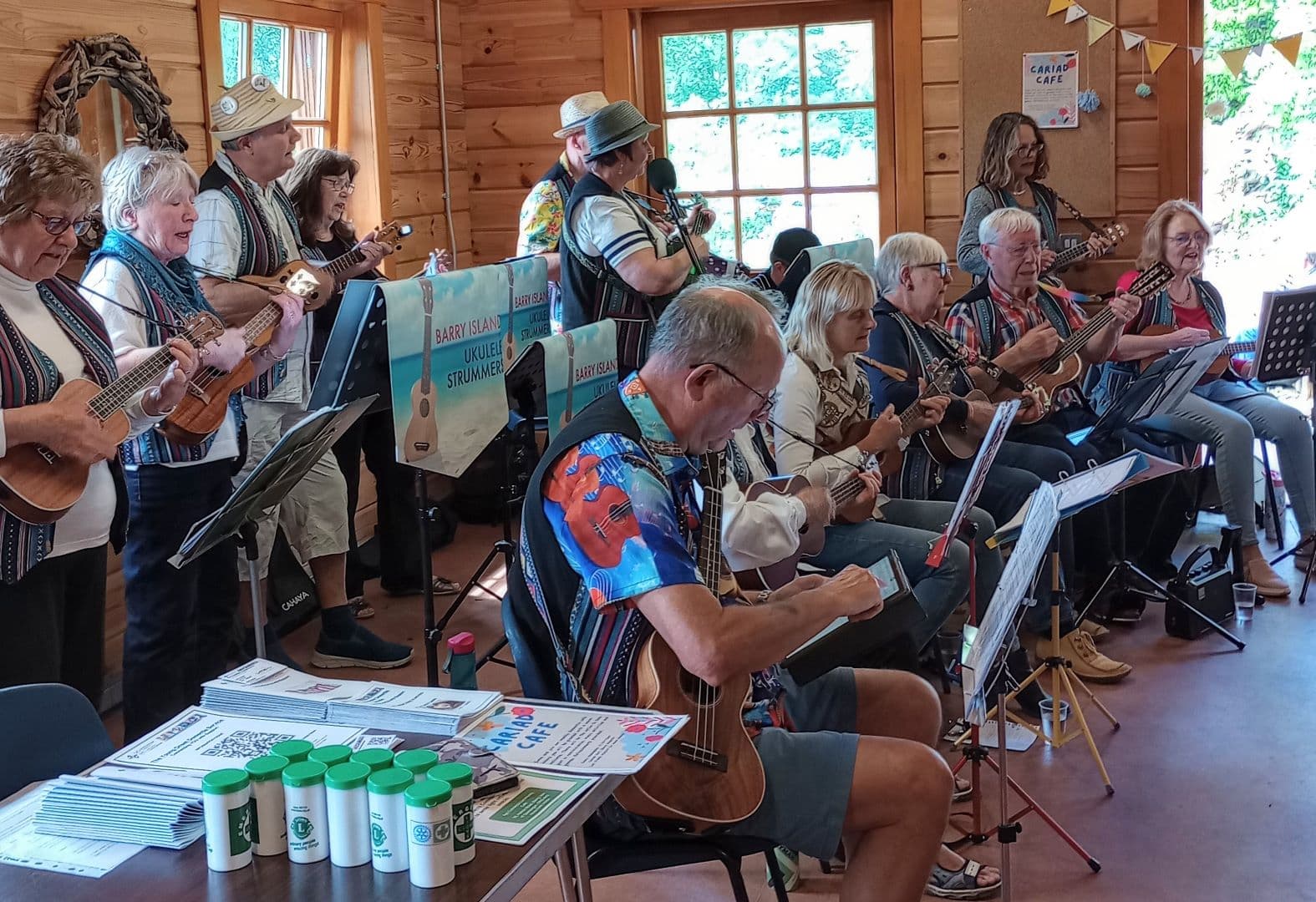 A group of people playing ukuleles indoors near a window, with a table of materials and a banner in the background. - Home Instead