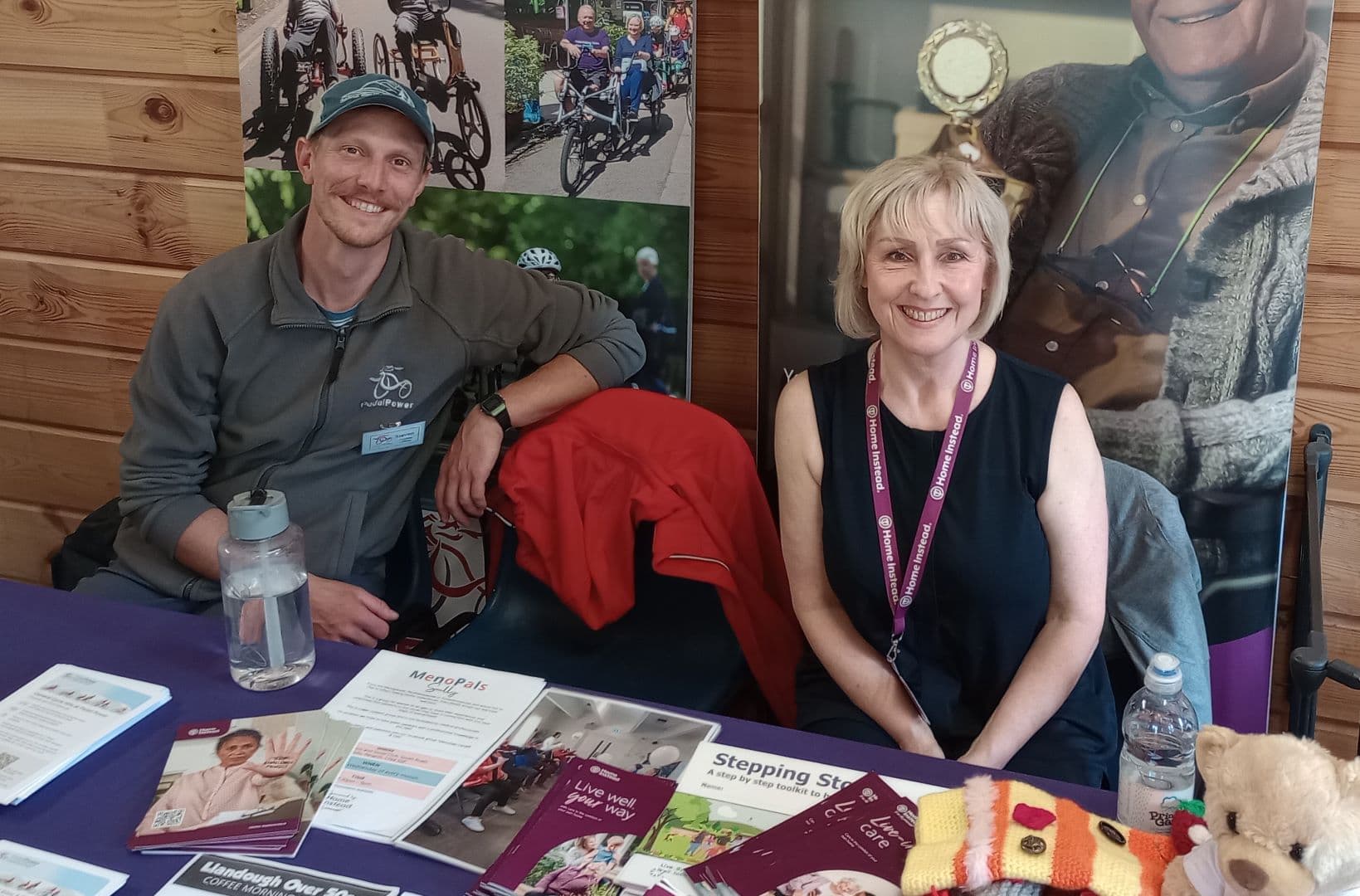 Two people sit at an information booth with pamphlets, wearing name badges and smiling at the camera. - Home Instead