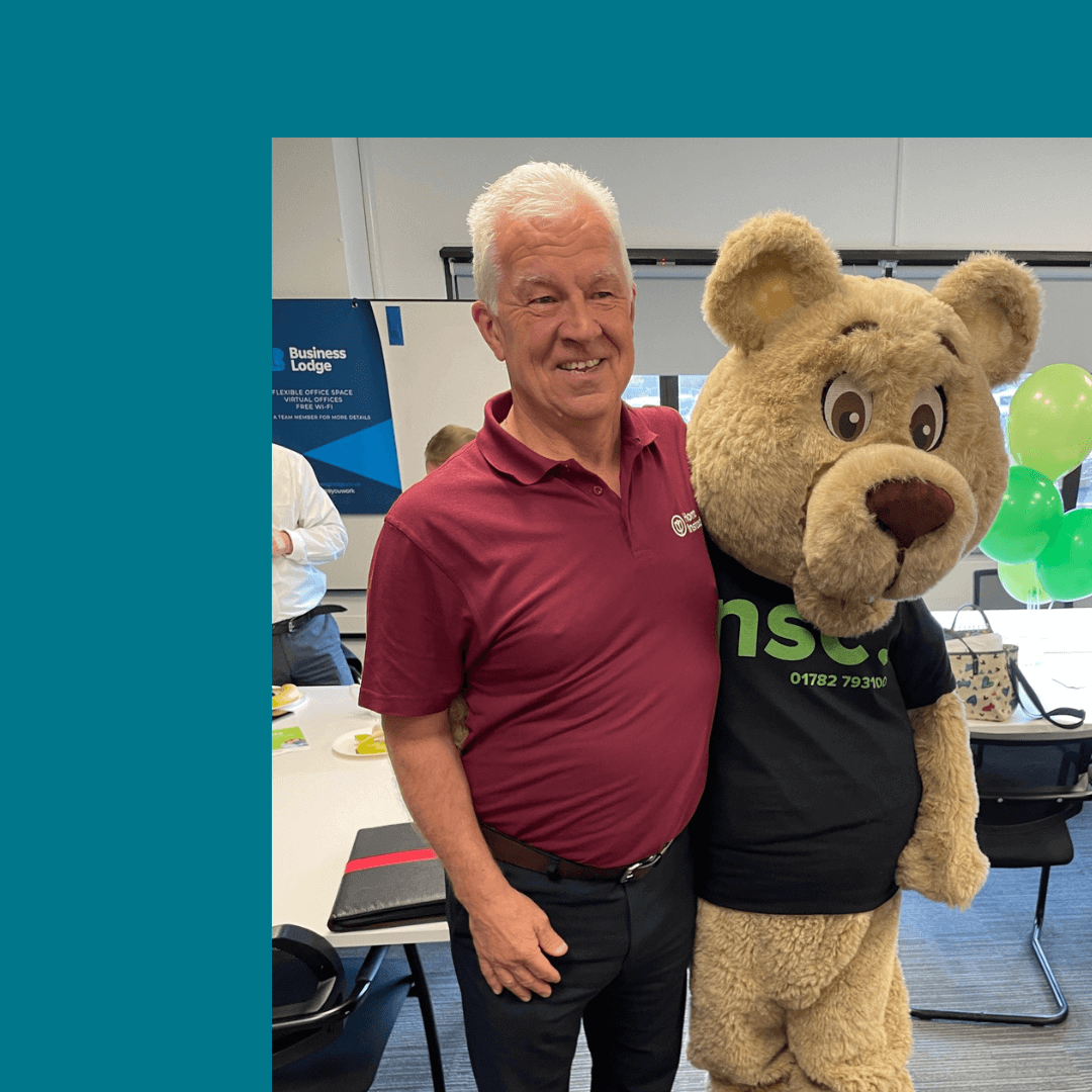 A smiling man in a maroon shirt stands next to a person in a bear costume at an indoor event. - Home Instead