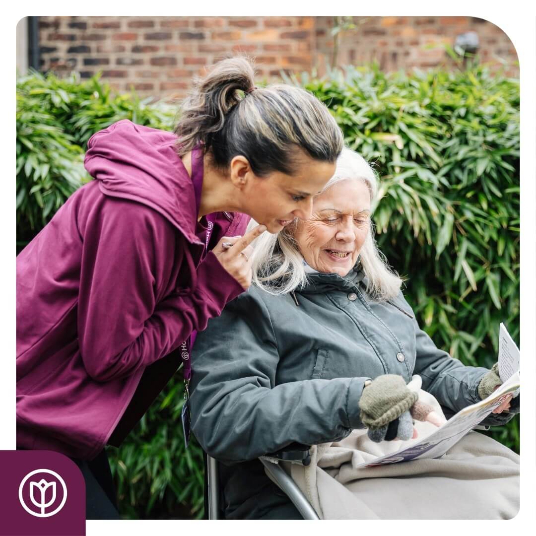 A caregiver in a maroon jacket interacts with an elderly woman in a wheelchair holding a newspaper. - Home Instead