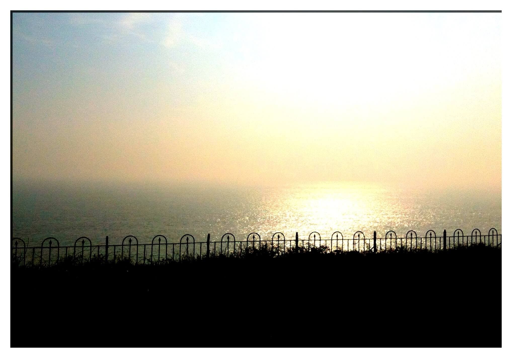 Silhouetted fence in front of a calm sea with sunlight reflecting off the water under a hazy morning sky. - Home Instead