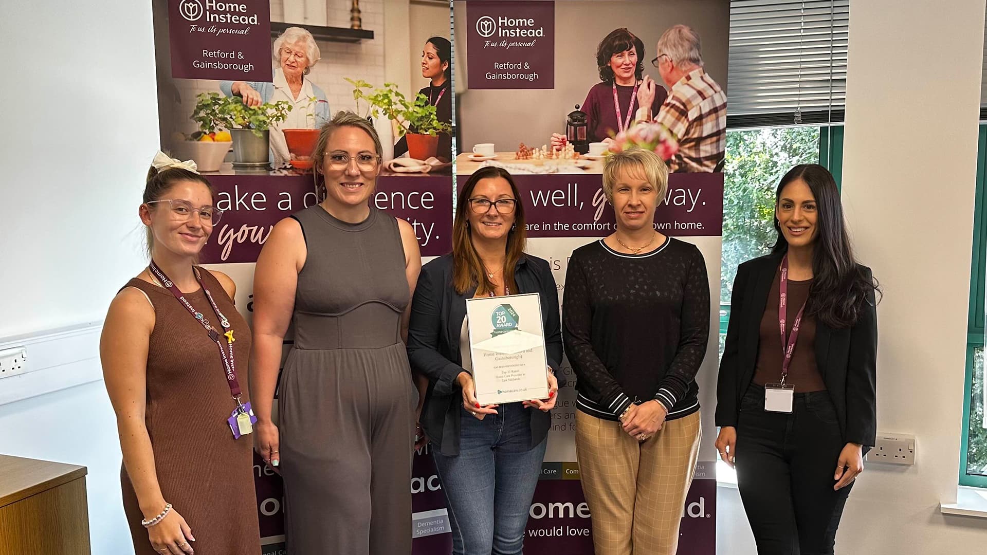 A group of five women standing together indoors, with one holding a framed certificate, smiling towards the camera. - Home Instead