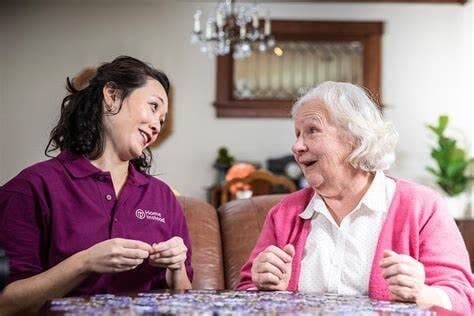 Two women, one younger in a purple shirt and one older in a pink cardigan, smiling at each other while seated indoors. - Home Instead