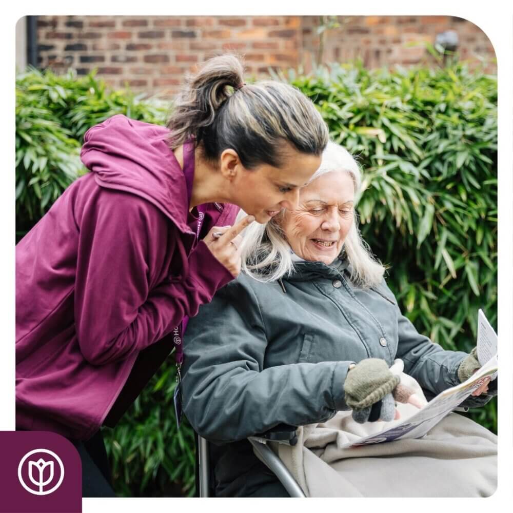 Caregiver in a purple hoodie leans in to talk to an elderly woman in a wheelchair holding a newspaper. - Home Instead