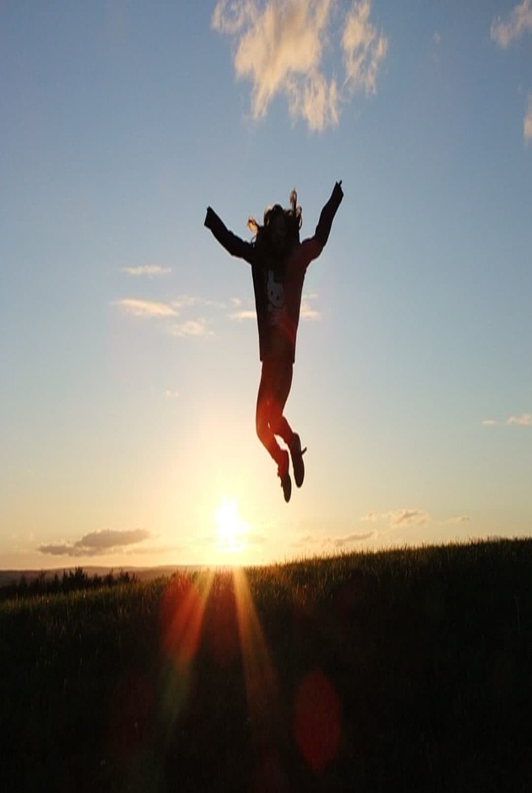 Silhouette of a person jumping joyfully against a sunset sky with clouds, above a grassy hill. - Home Instead