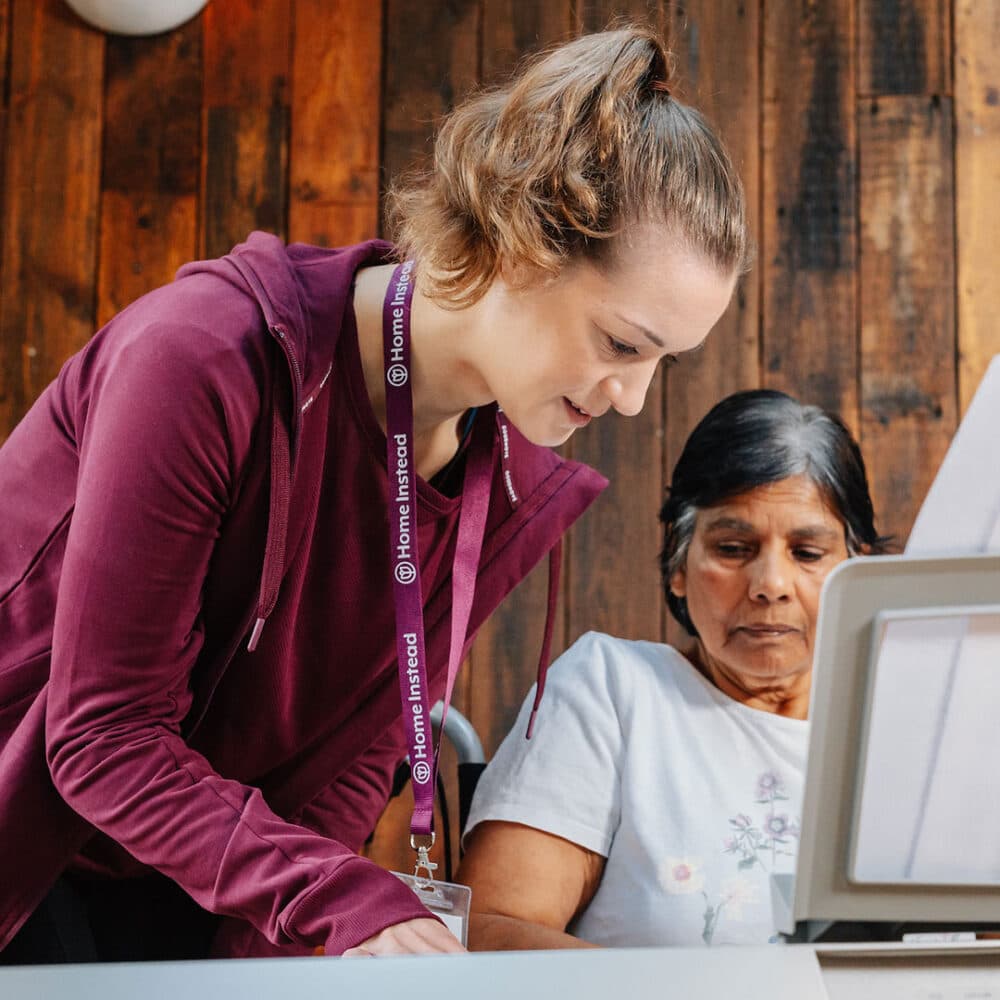 A caregiver in a maroon jacket assists an elderly woman using a laptop. They are in a wooden-panelled room. - Home Instead