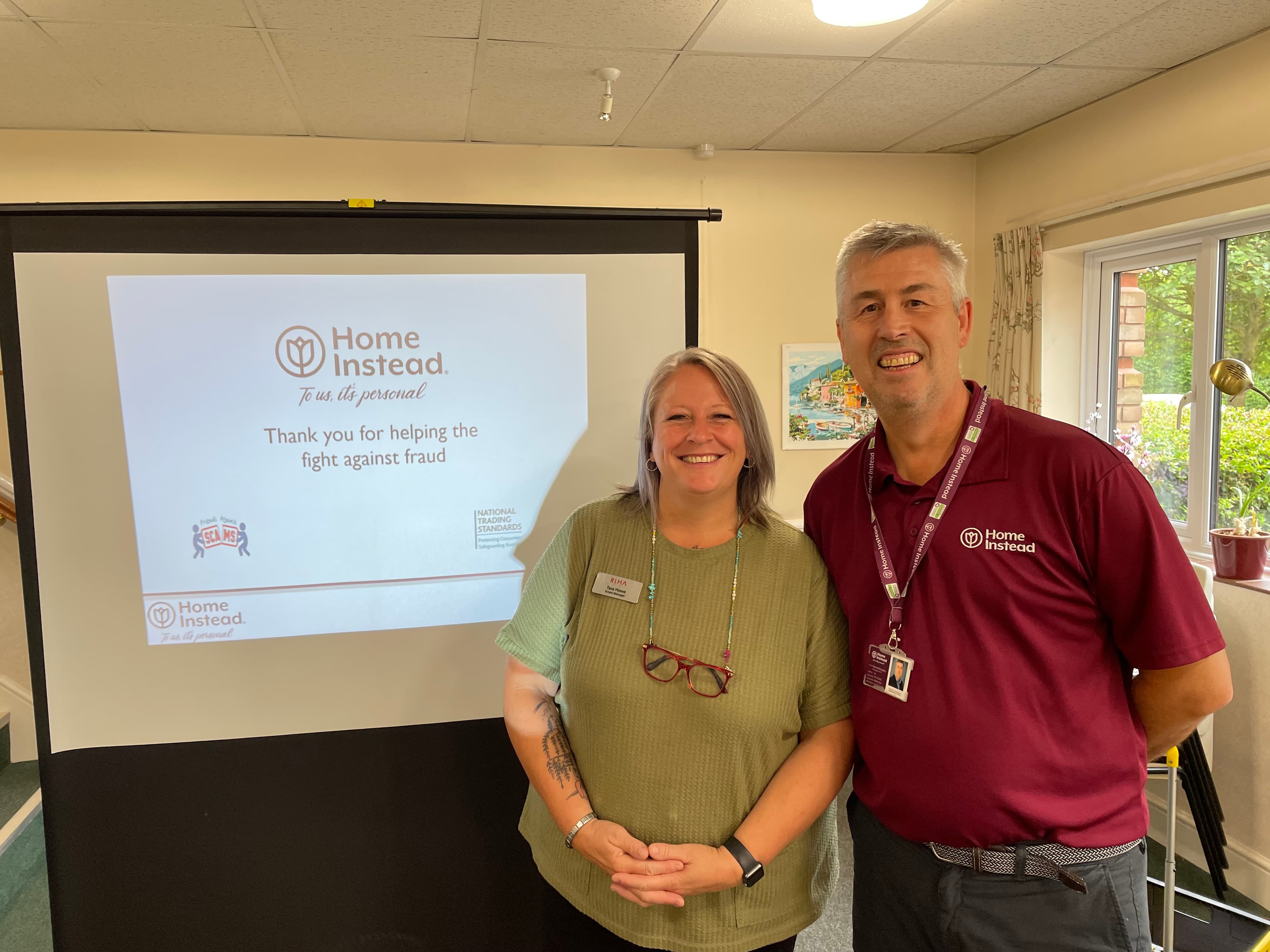 Two people smiling and standing in front of a presentation slide about fighting fraud, with "Home Instead" branding visible. - Home Instead