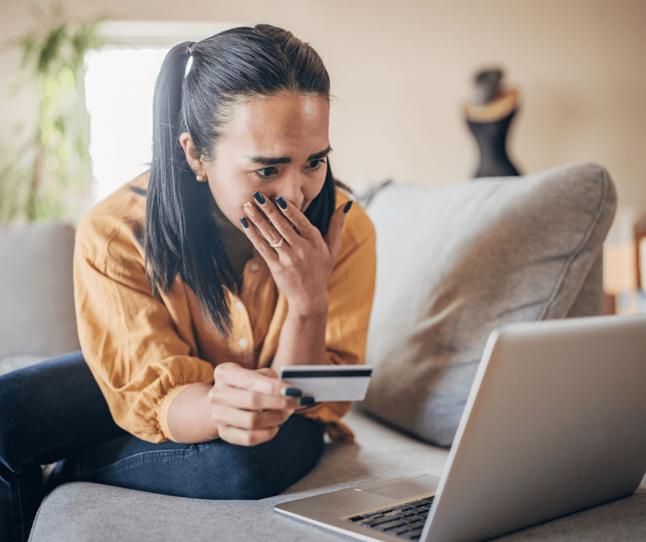 Woman holding a credit card, looking surprised at a laptop screen, sitting on a couch in a living room. - Home Instead