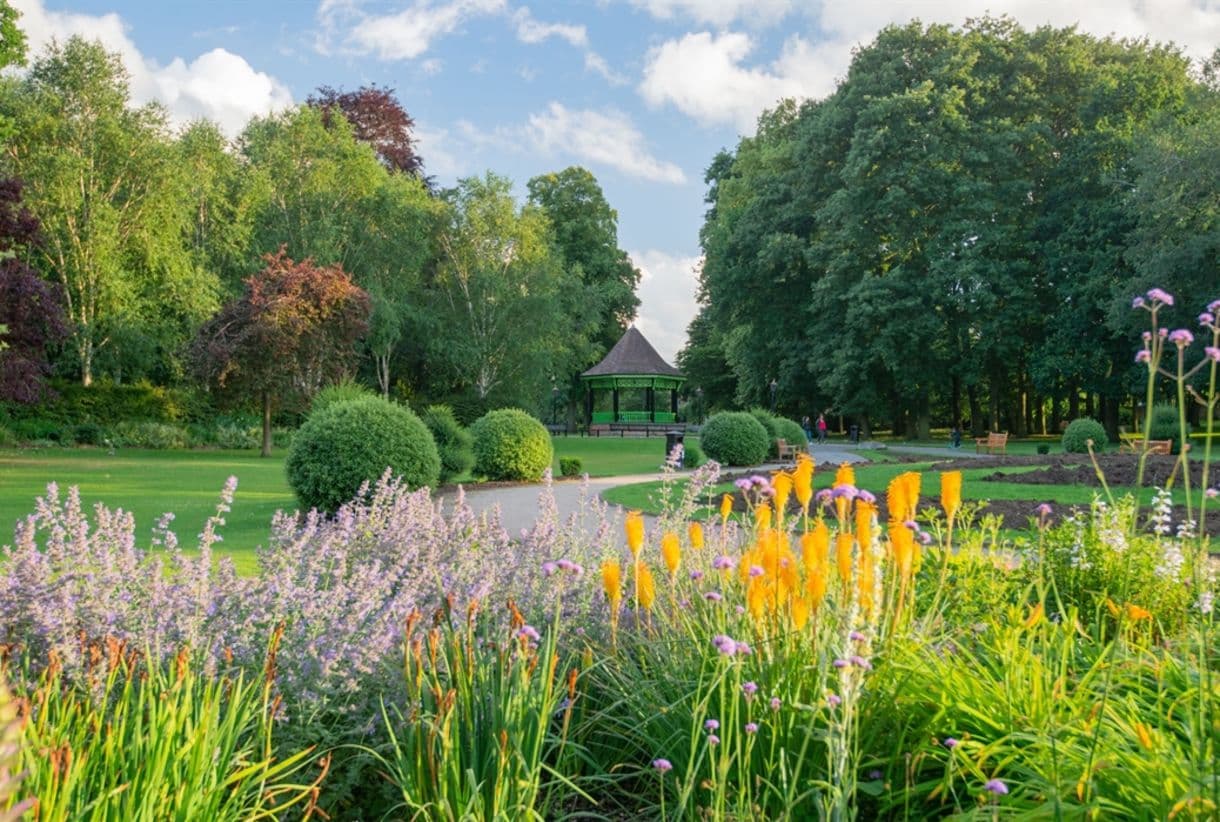 A beautiful park with vibrant flowers, a paved pathway, green trees, and a gazebo under a partly cloudy sky. - Home Instead