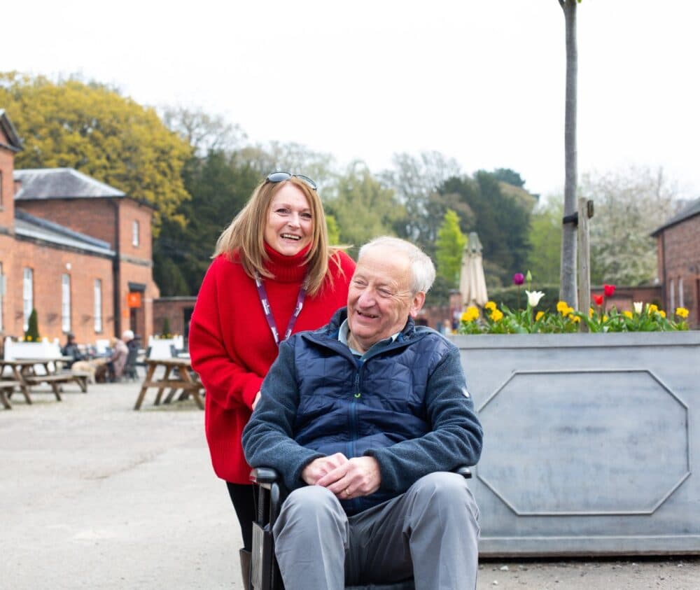 A woman in a red sweater smiles while standing behind an elderly man in a wheelchair outdoors. - Home Instead