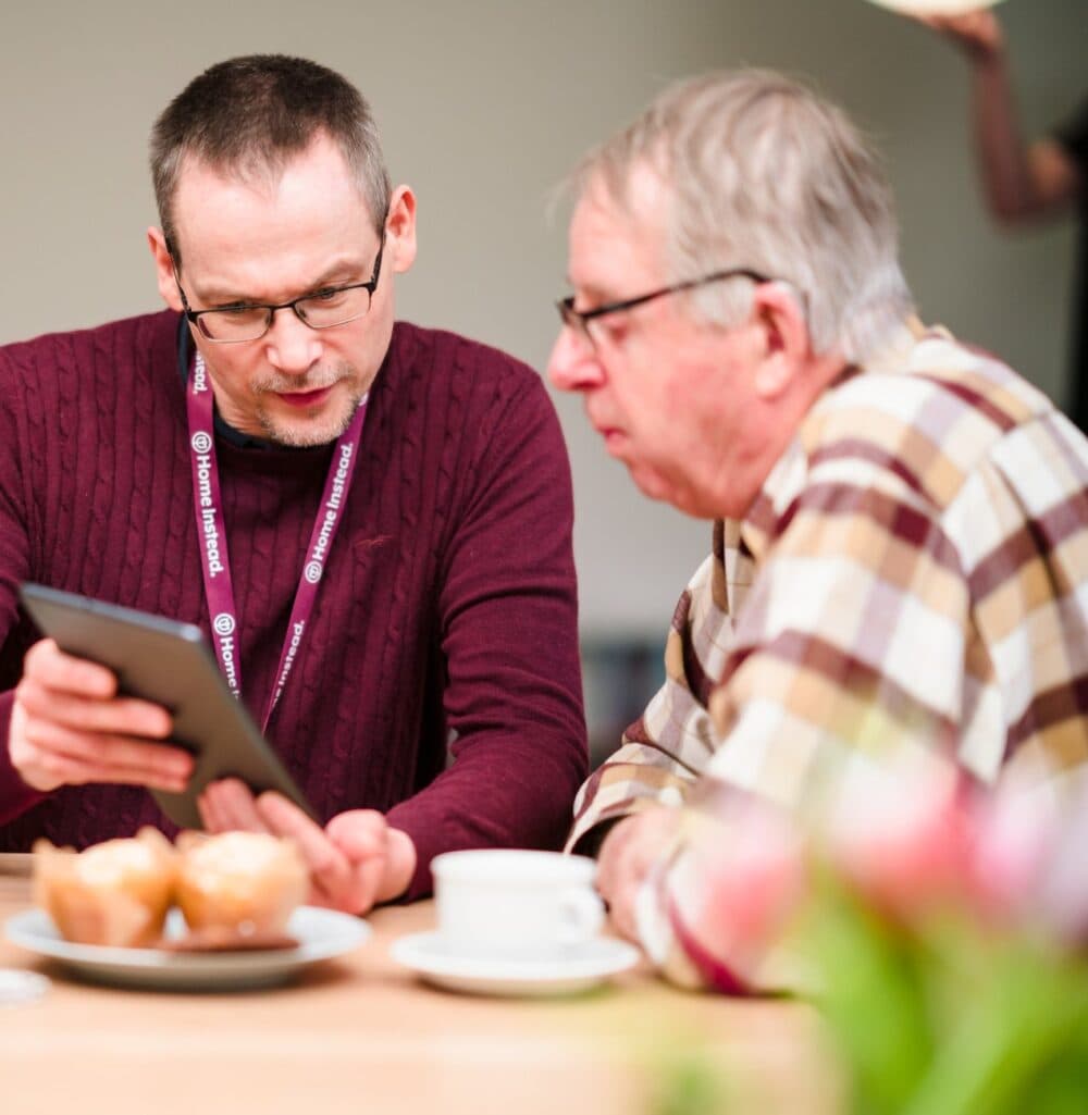 Two men having a conversation at a table, one showing something on a tablet to the other. Coffee and muffins on the table. - Home Instead