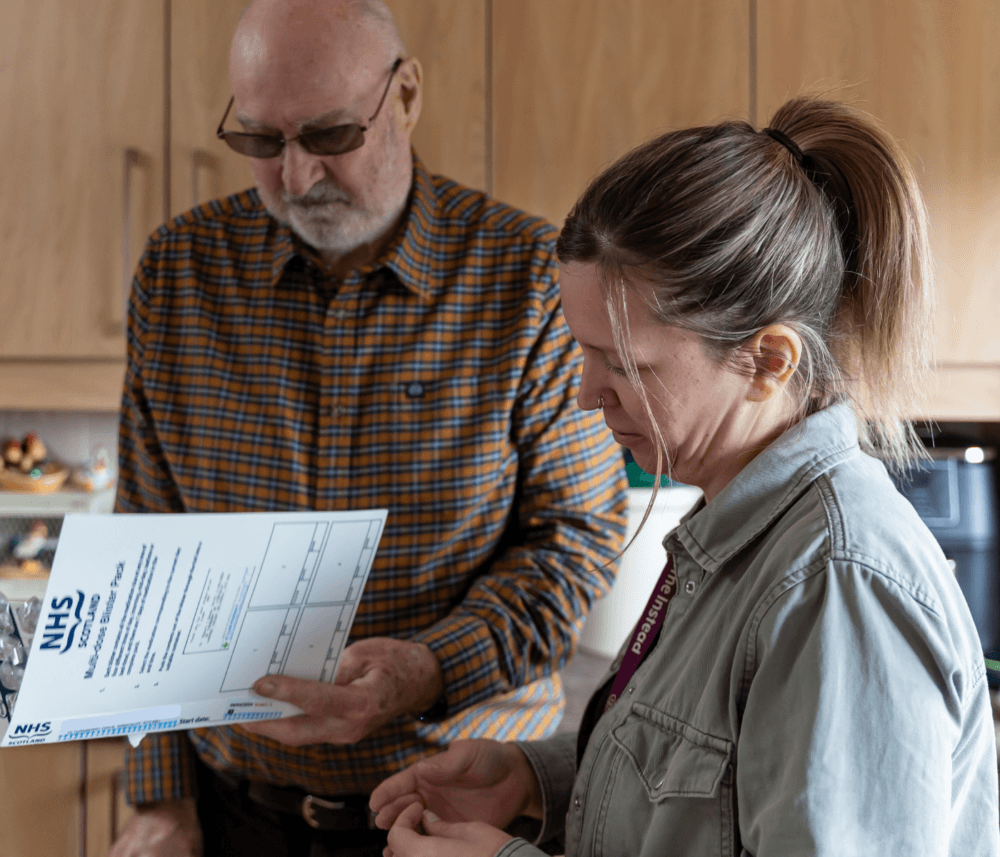 A man and a woman review an NHS document together in a kitchen with wooden cabinets in the background. - Home Instead