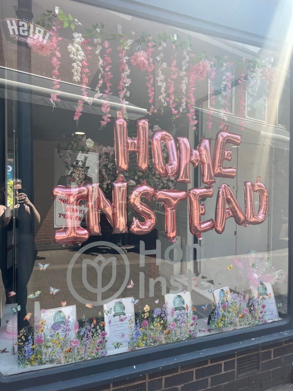 A storefront window with pink balloon letters spelling "HOME INSTEAD" amid floral decorations and gift items. - Home Instead