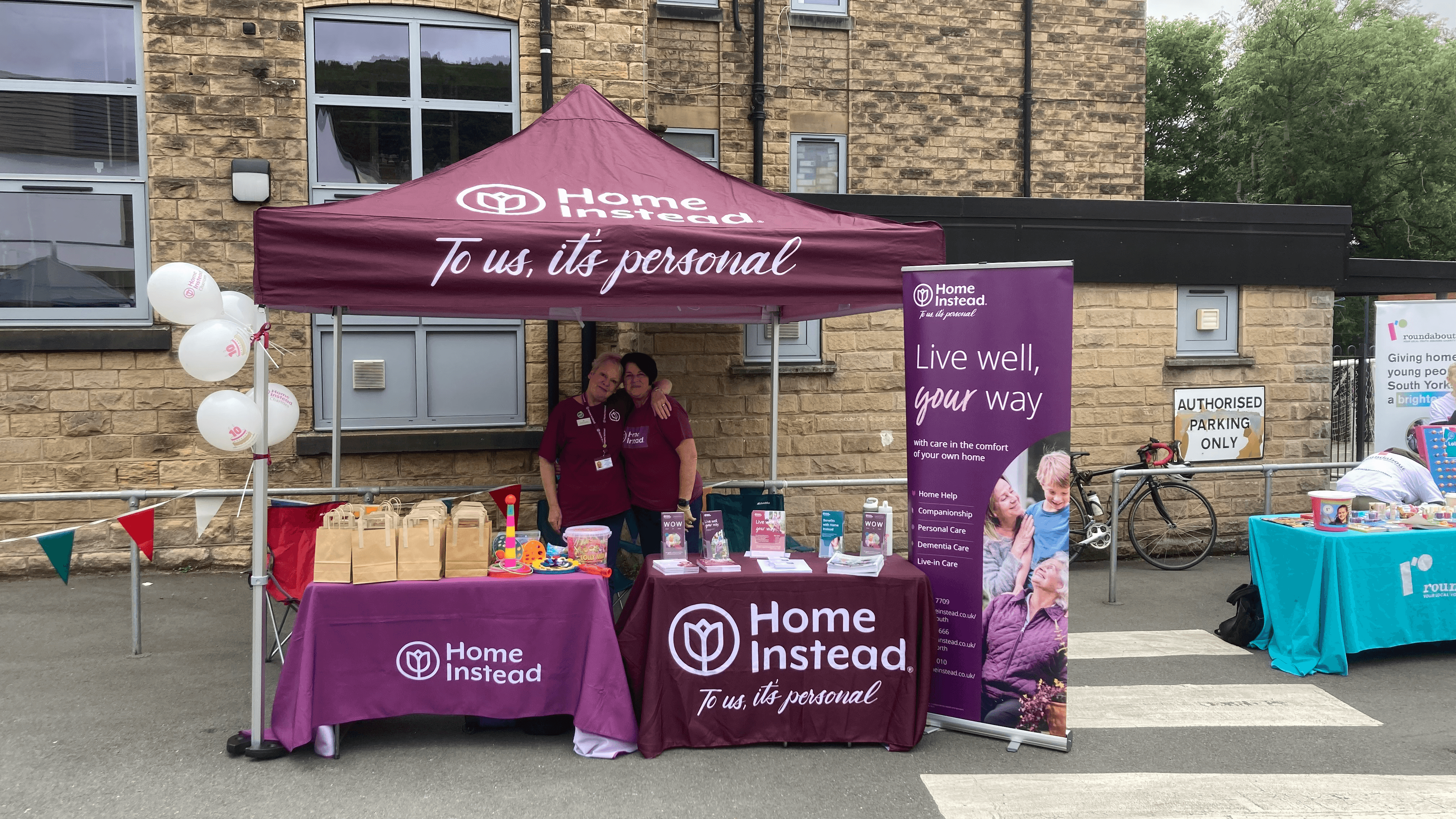 A Home Instead booth with informational displays and staff under a purple tent at an outdoor event near a stone building. - Home Instead