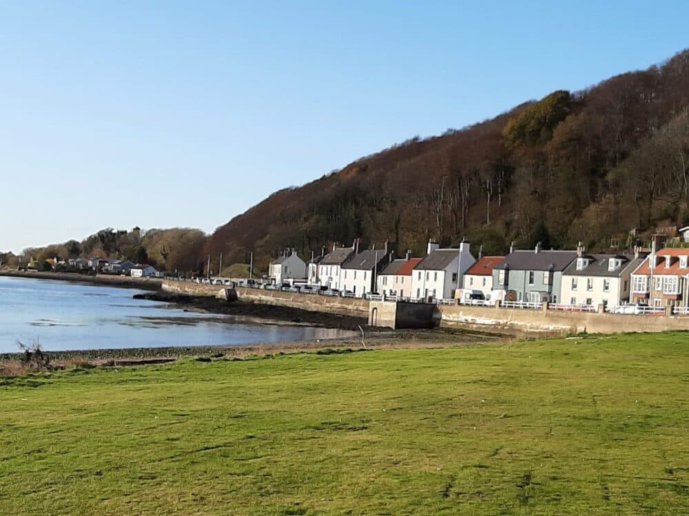 Row of colorful houses along a waterfront with a grassy field in the foreground and a hill in the background. - Home Instead