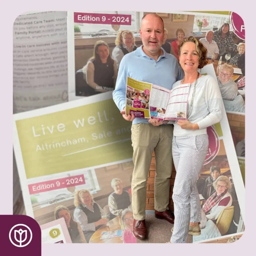 Two people holding an open magazine, standing in front of a large backdrop featuring images of a community setting. - Home Instead