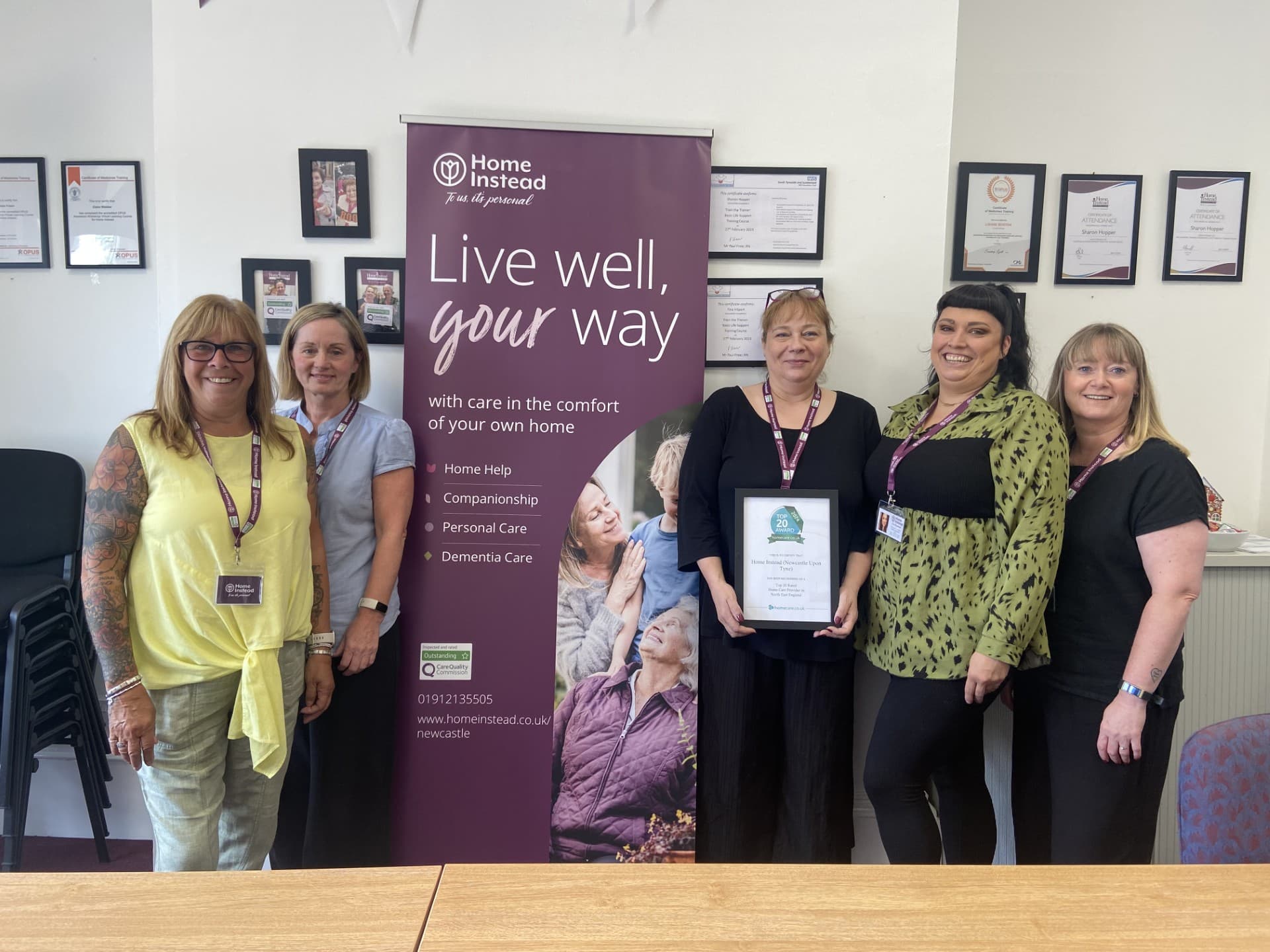 A group of five people standing in front of a Home Instead banner, one of whom is holding a framed certificate. - Home Instead