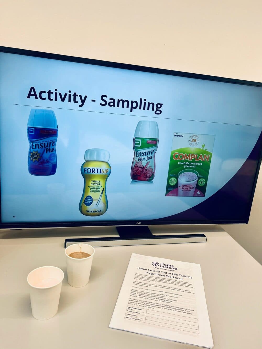 A screen displays different nutritional drinks, with two paper cups and a document on a table in the foreground. - Home Instead