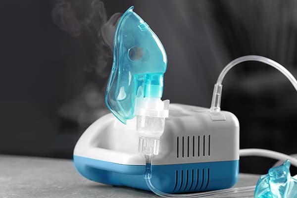 A nebulizer with a blue mask attached, emitting vapor, on a gray surface against a dark background. - Home Instead