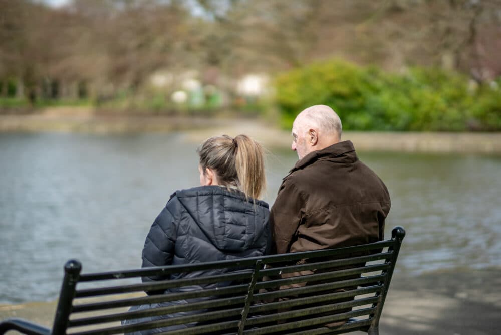 Two people, seen from behind, sit on a park bench by a lake on a cloudy day. - Home Instead
