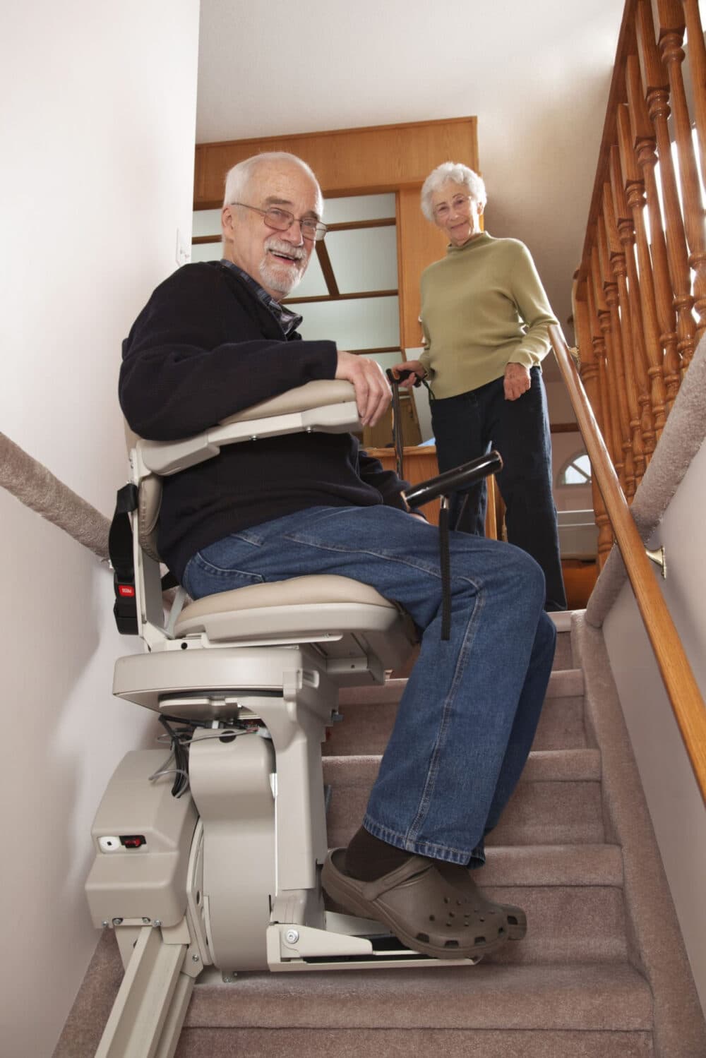 An elderly man using a stair lift on a staircase, while a smiling elderly woman stands at the top of the stairs. - Home Instead