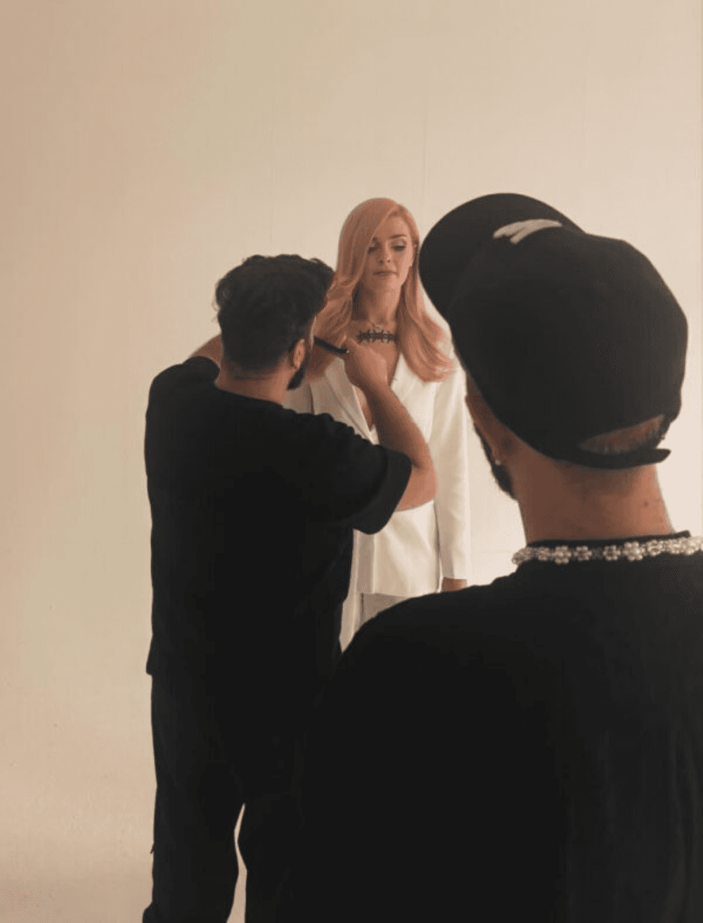 A stylist adjusts a necklace on a person with long pink hair, wearing a white outfit, with another person observing. - Home Instead