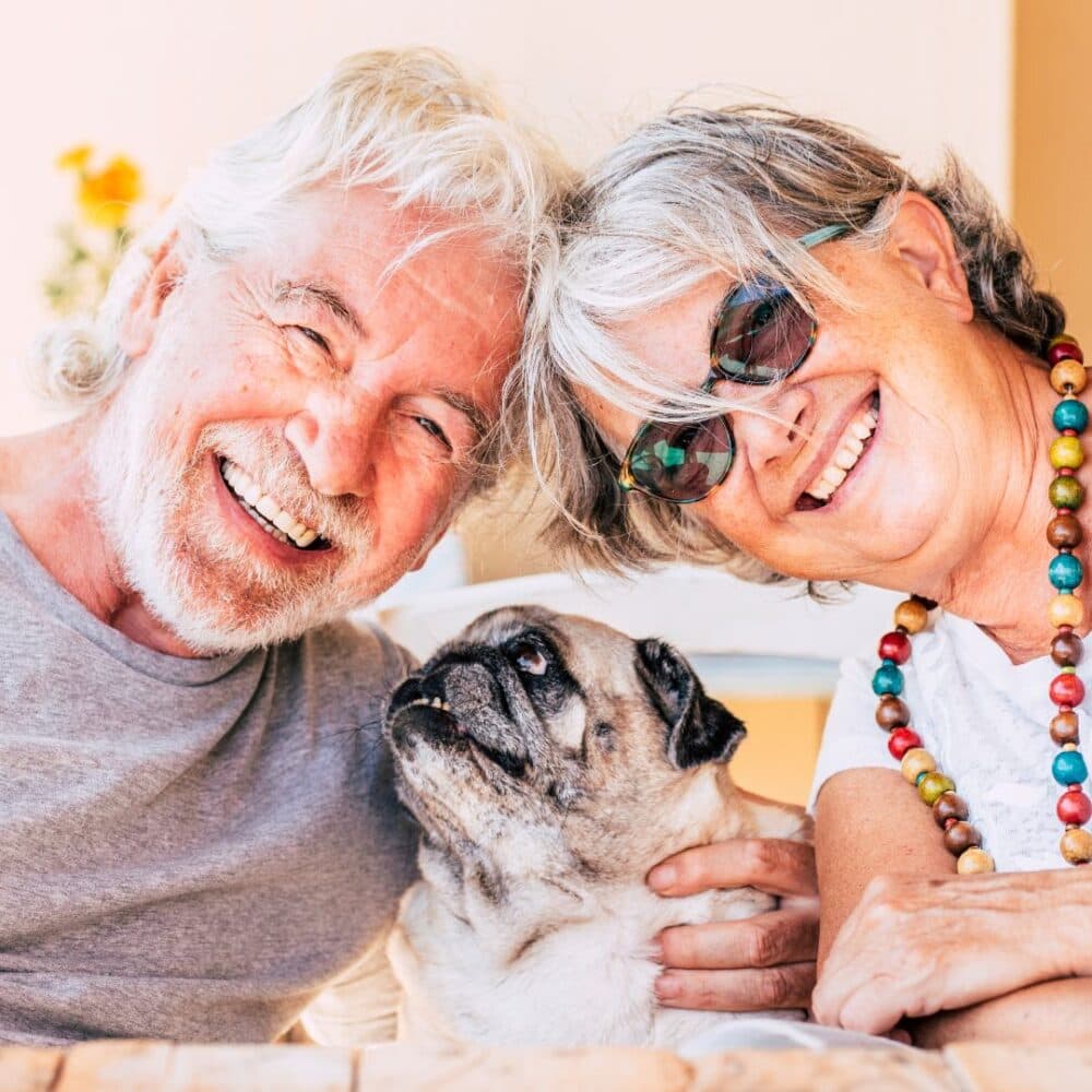 Smiling elderly couple with gray hair hold a pug, sitting closely and enjoying their time together. - Home Instead