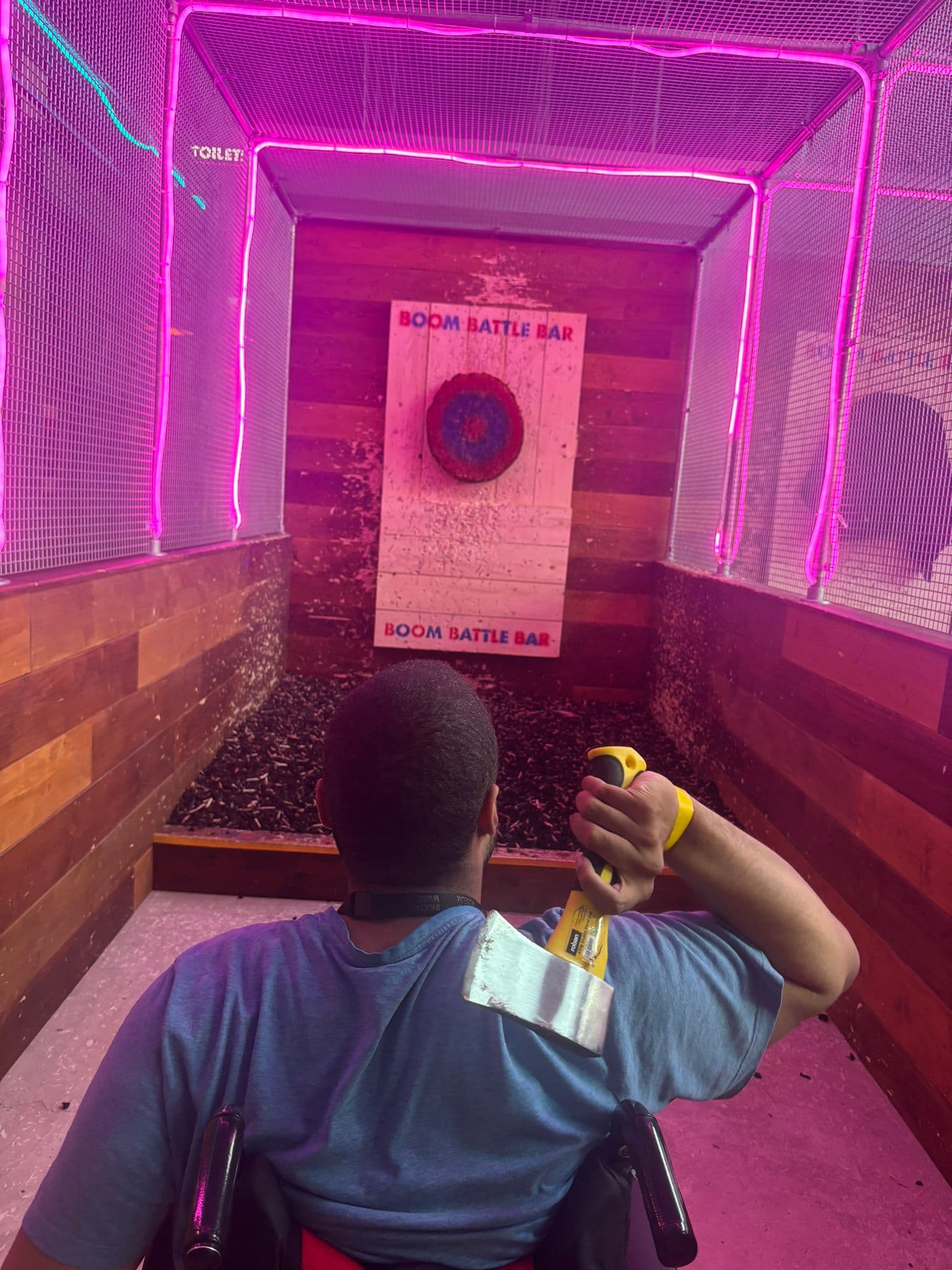 Person in a wheelchair throwing an axe at a target in an axe-throwing venue with neon lights and wooden walls. - Home Instead