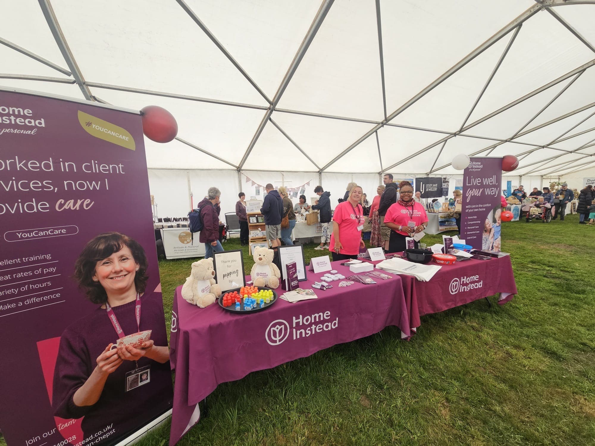 Booth under a large tent showcasing "Home Instead" with staff, informational materials, and promotional items on tables. - Home Instead