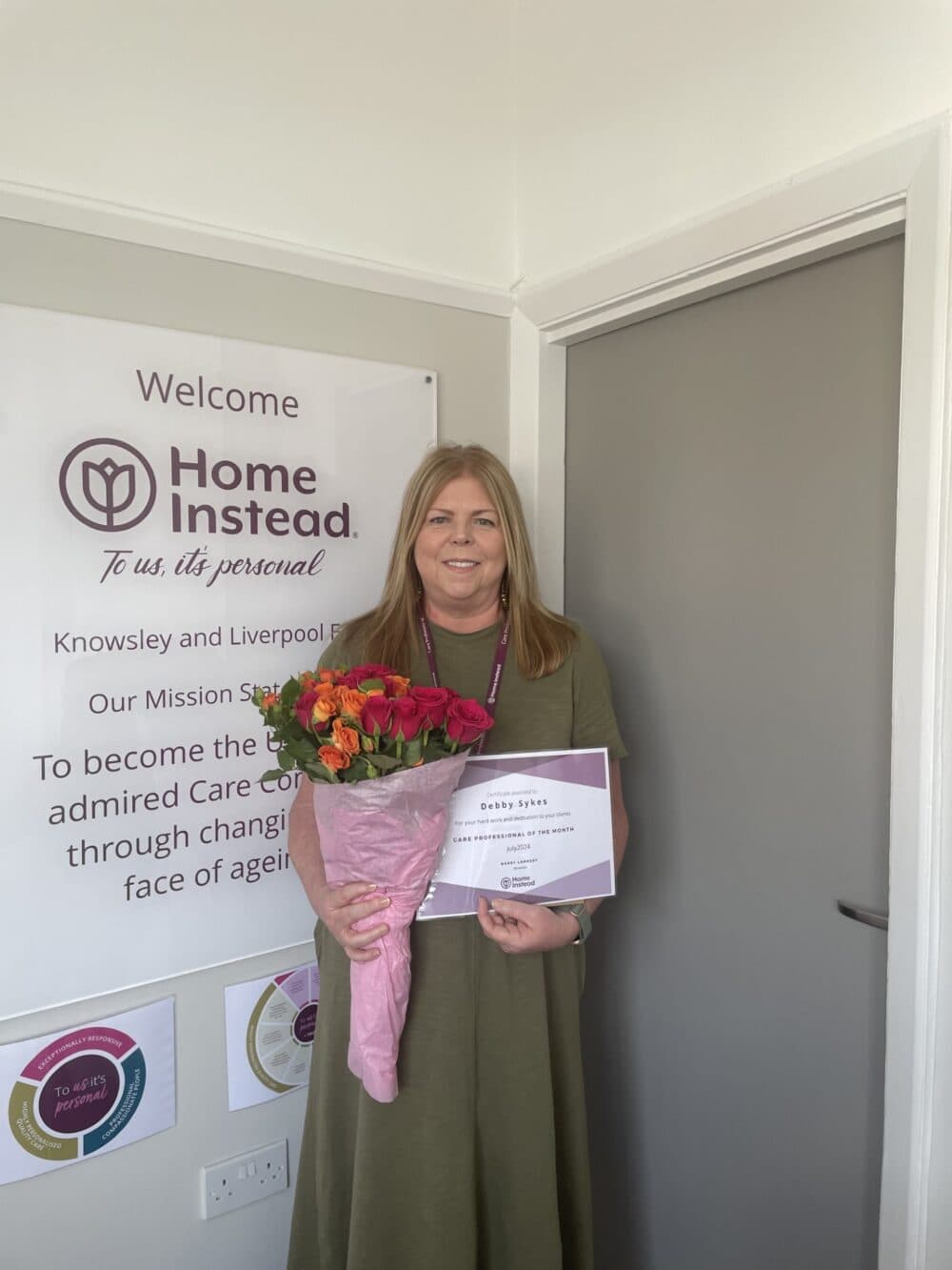 Woman holding a bouquet of flowers and a certificate, standing next to a Home Instead sign on the wall. - Home Instead