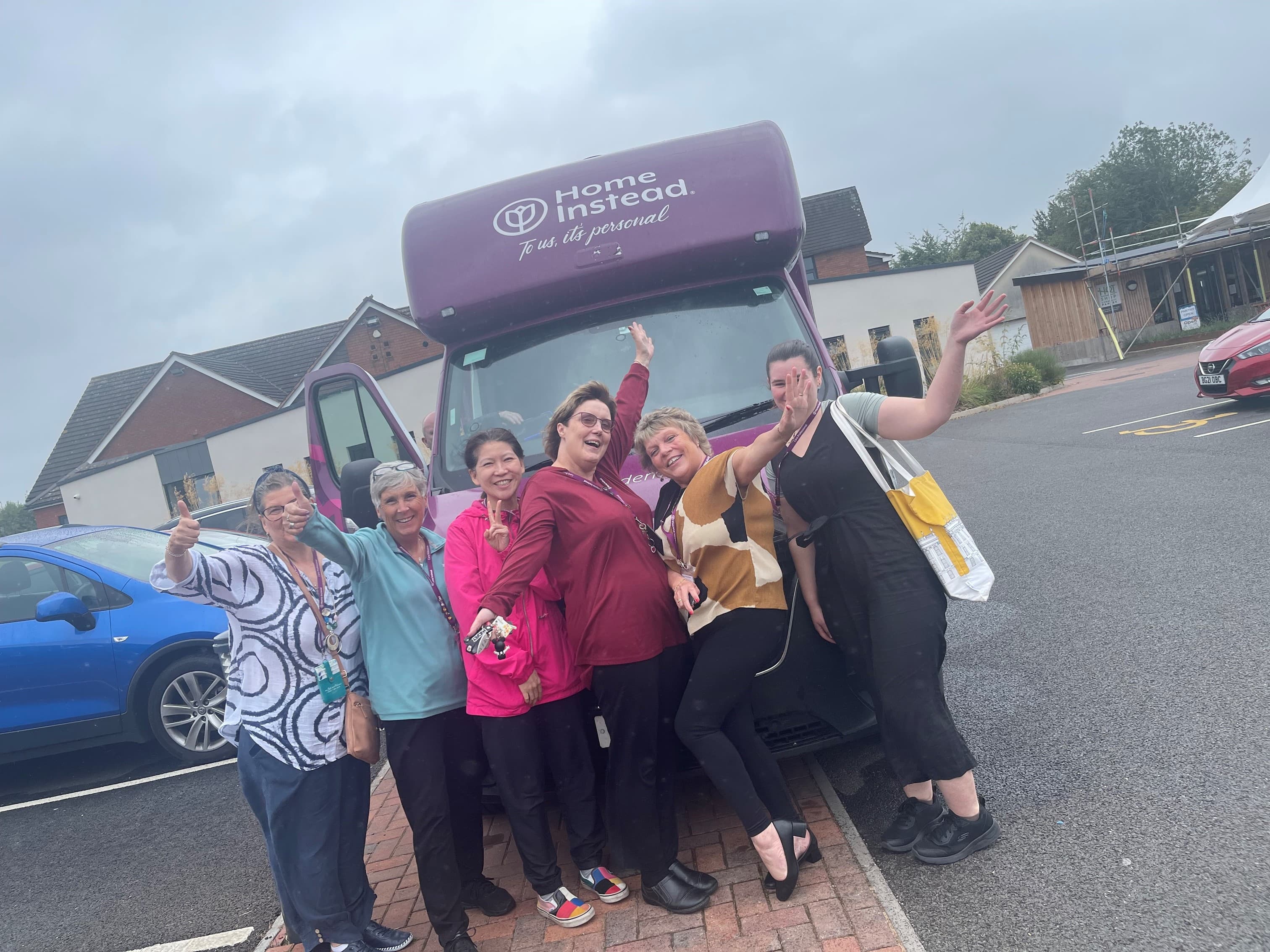 Six Home Instead Care Professionals, smiling and waving standing in front of the Dementia Tour Bus - Home Instead