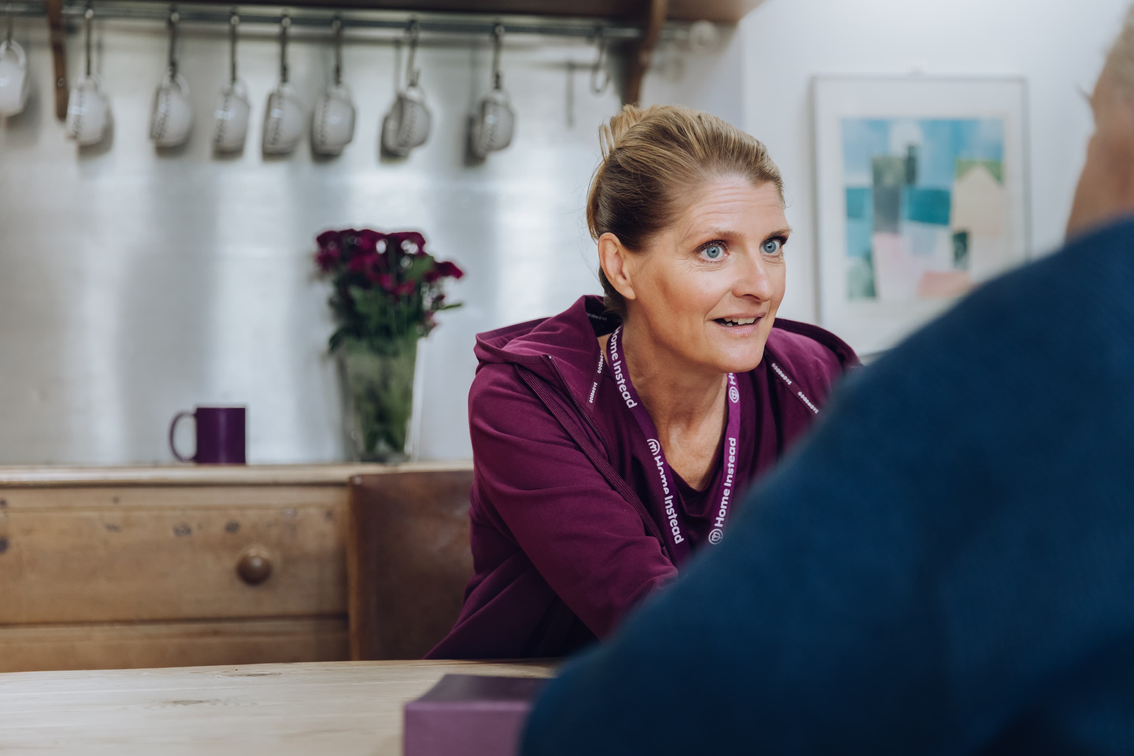 A woman in a purple uniform talks to a person. A mug and flowers are on a wooden table in the background. - Home Instead