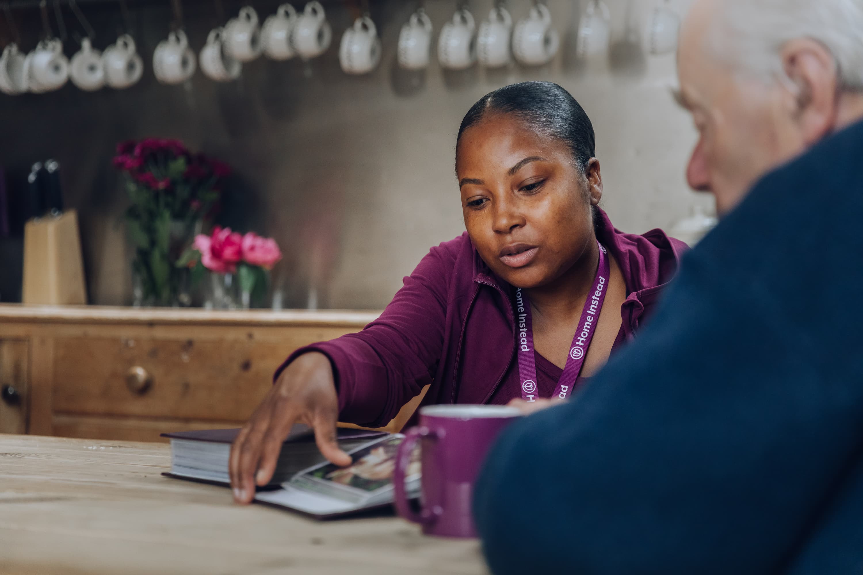 A woman shows a book to an older man at a kitchen table with mugs and flowers. - Home Instead
