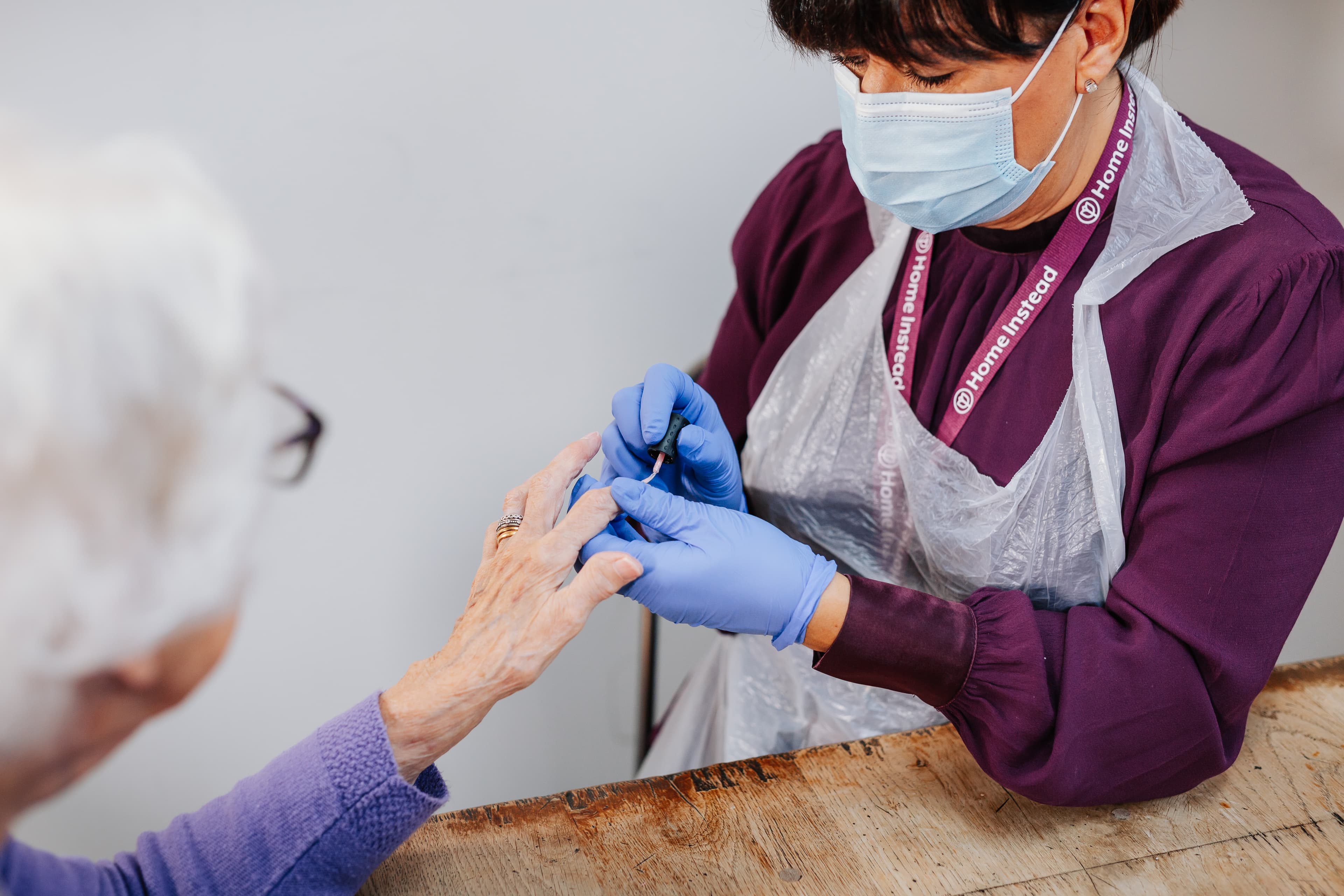 A caregiver in gloves and a mask assists an elderly person with a finger splint at a table. - Home Instead