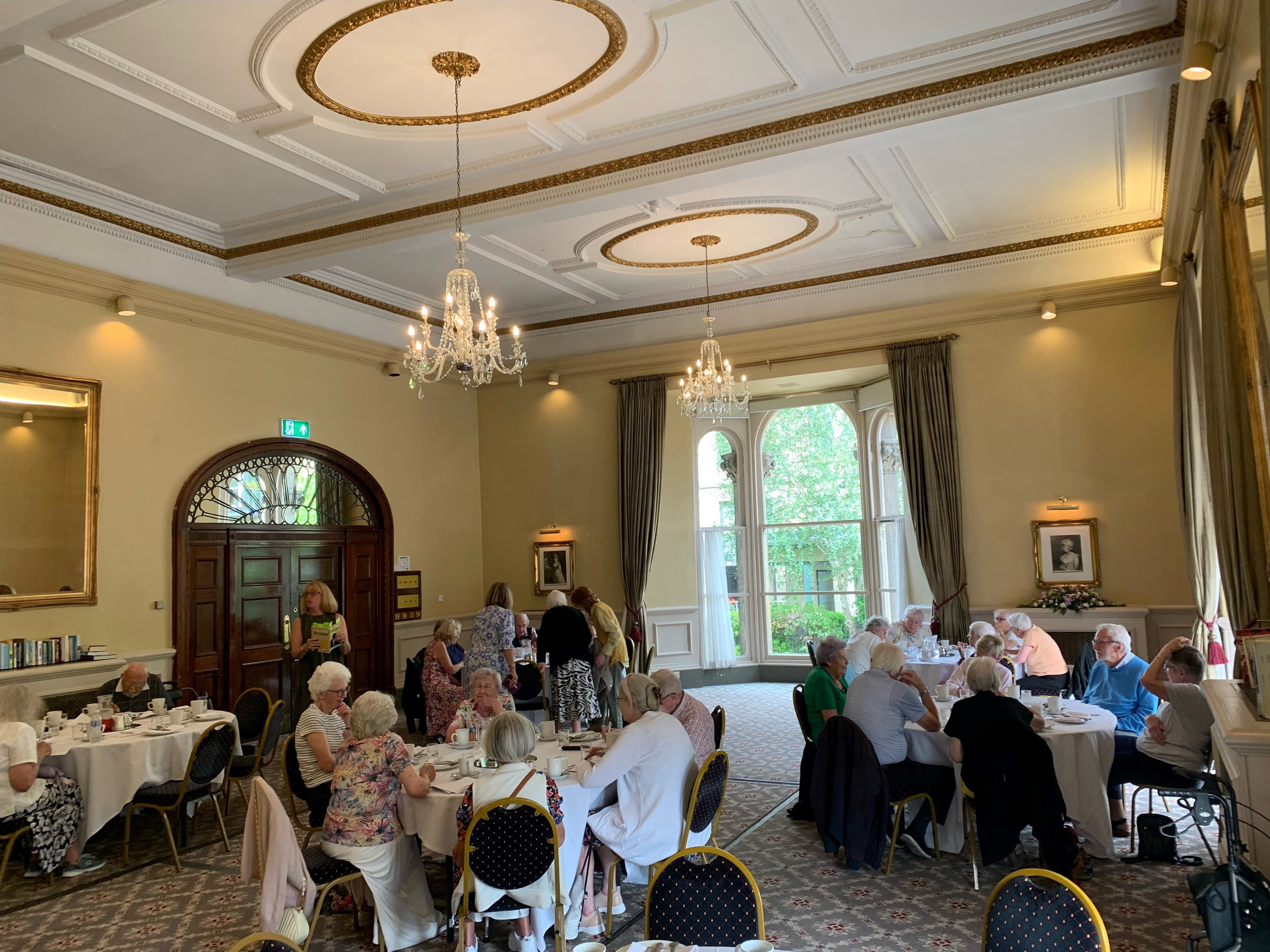 A group of elderly people dining in an elegant room with chandeliers, high ceilings, and large windows. - Home Instead