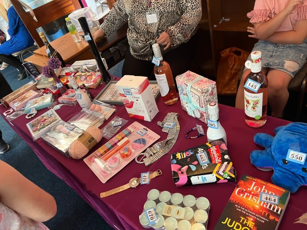 A table with various items for sale, including skincare products, books, wine, and accessories, each labeled with a price tag. - Home Instead