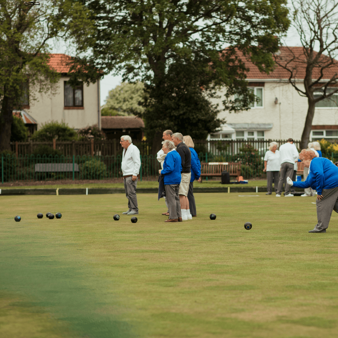 People playing lawn bowls on a green field with houses and trees in the background. - Home Instead