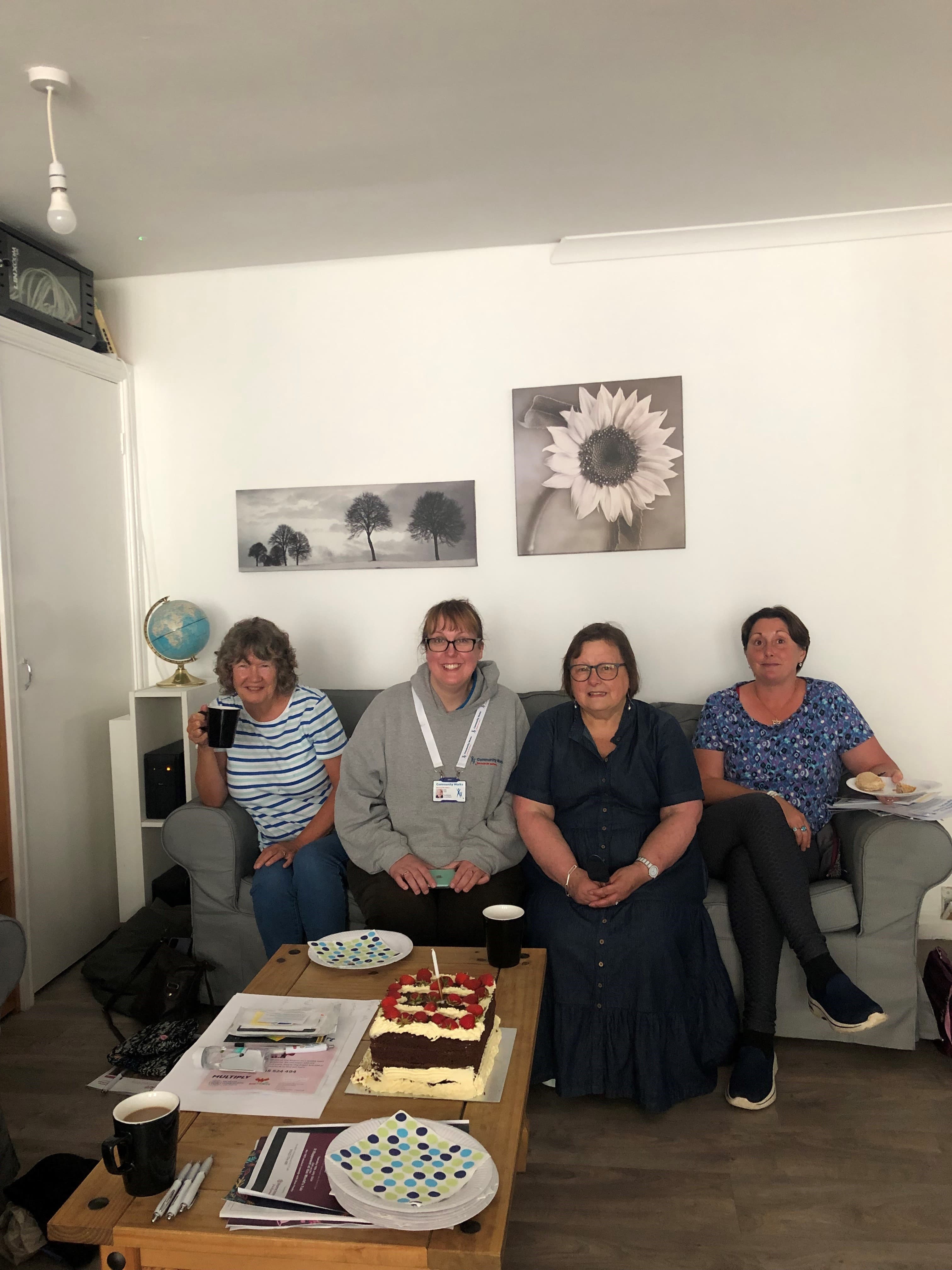 Four women sit in a living room around a table with a cake and plates, smiling at the camera. - Home Instead