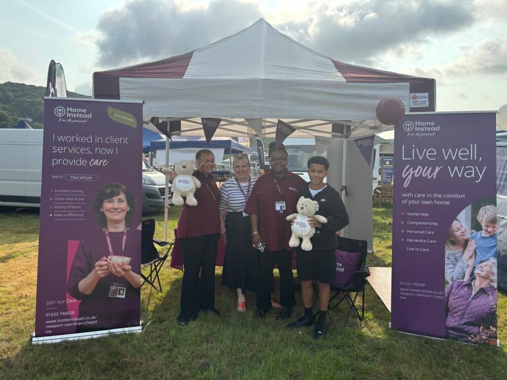Four people smiling, standing in front of a booth with banners promoting in-home care services, holding stuffed teddy bears. - Home Instead