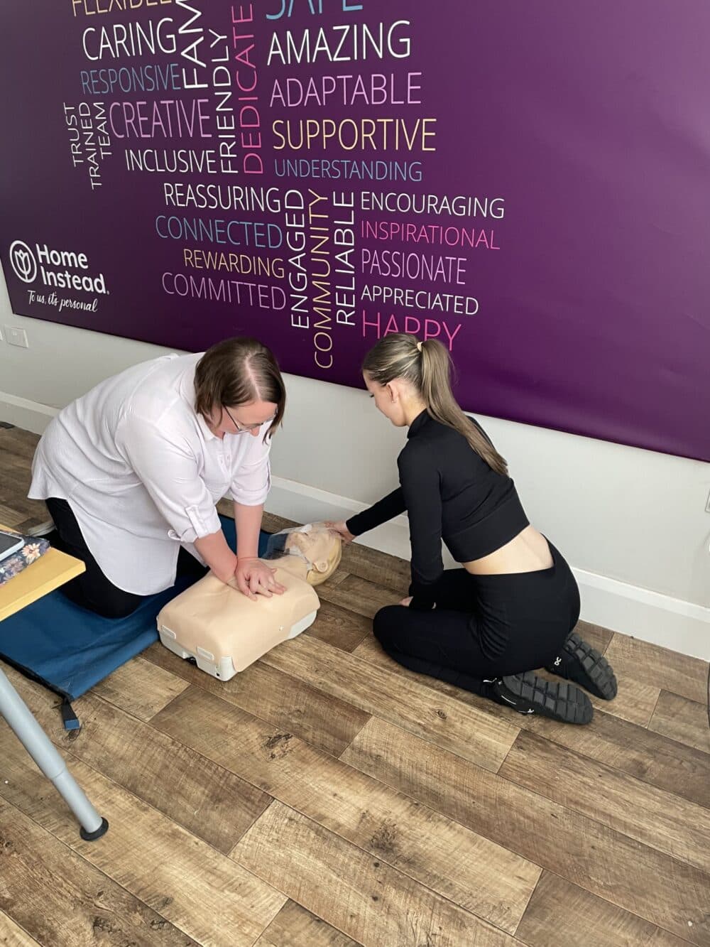 Two women practicing CPR on a dummy; one compressing the chest, the other observing, in a room with motivational words on wall. - Home Instead