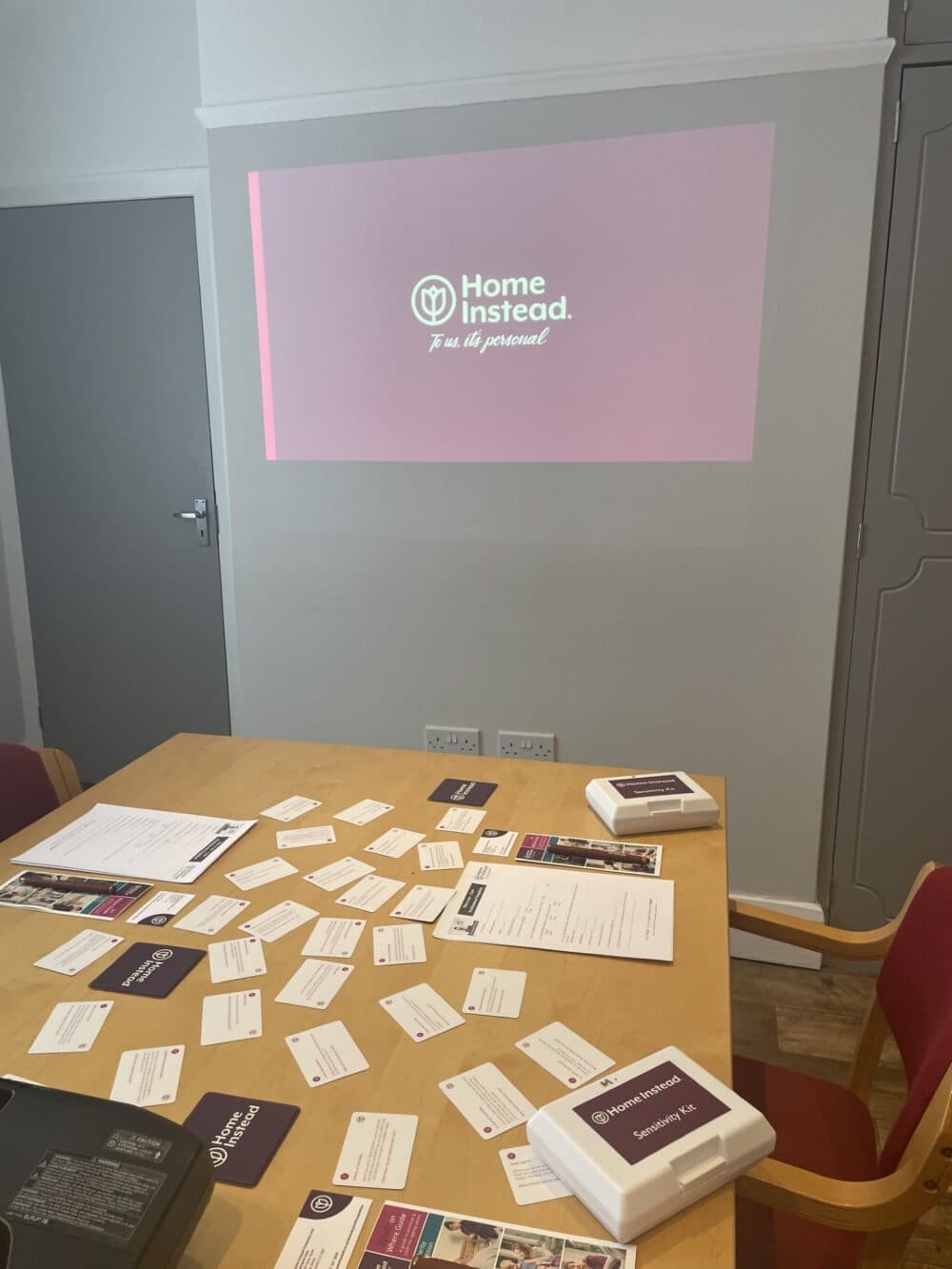 A conference room with a table covered in papers and materials, and a "Home Instead" presentation projected on the wall. - Home Instead