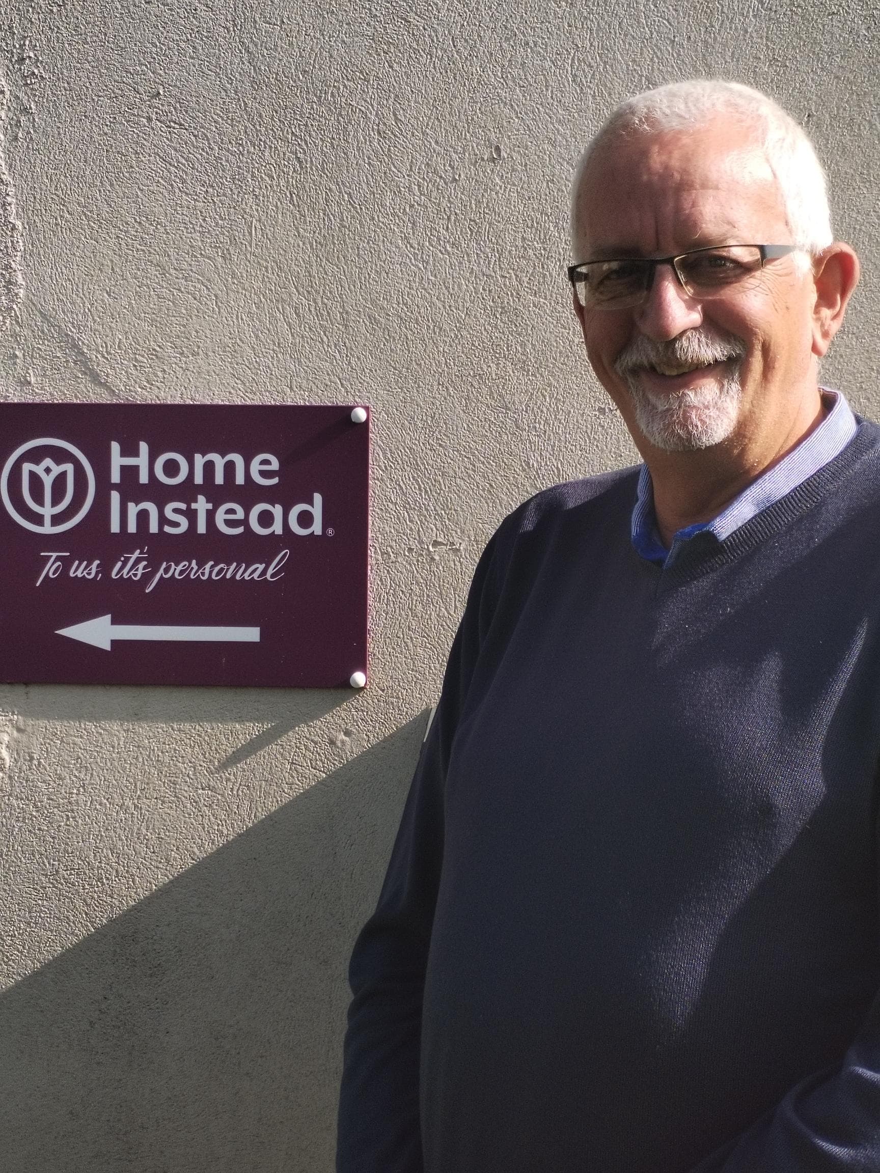Smiling man standing next to a "Home Instead" sign on a sunny day. - Home Instead