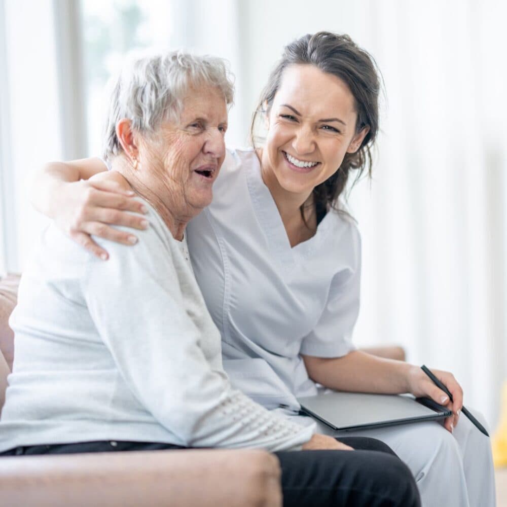 Nurse smiling and embracing a seated elderly woman, both looking happy in a bright room. - Home Instead