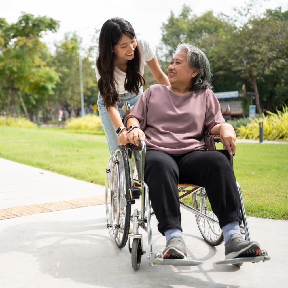 A woman smiles and pushes an elderly woman in a wheelchair along a sunny park pathway. - Home Instead