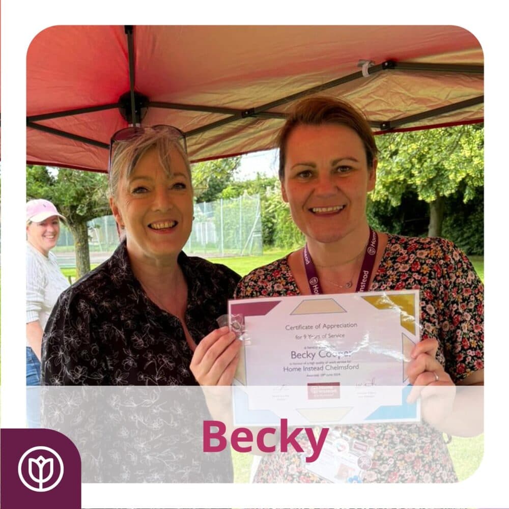 Two women smiling, with one holding a certificate of appreciation under a canopy. Trees are visible in the background. - Home Instead
