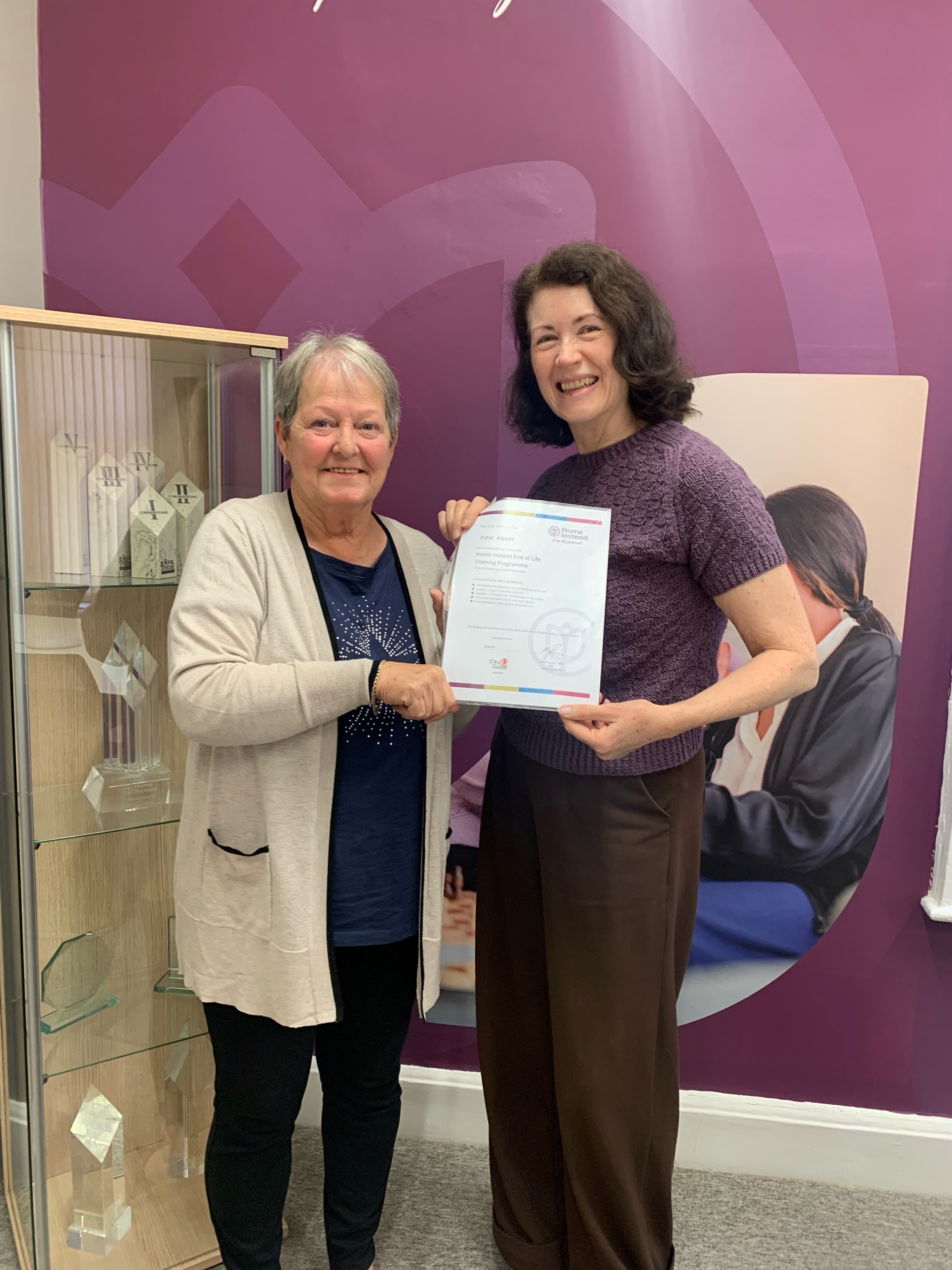 Two women smiling and holding a certificate in front of a purple wall and display case with awards. - Home Instead