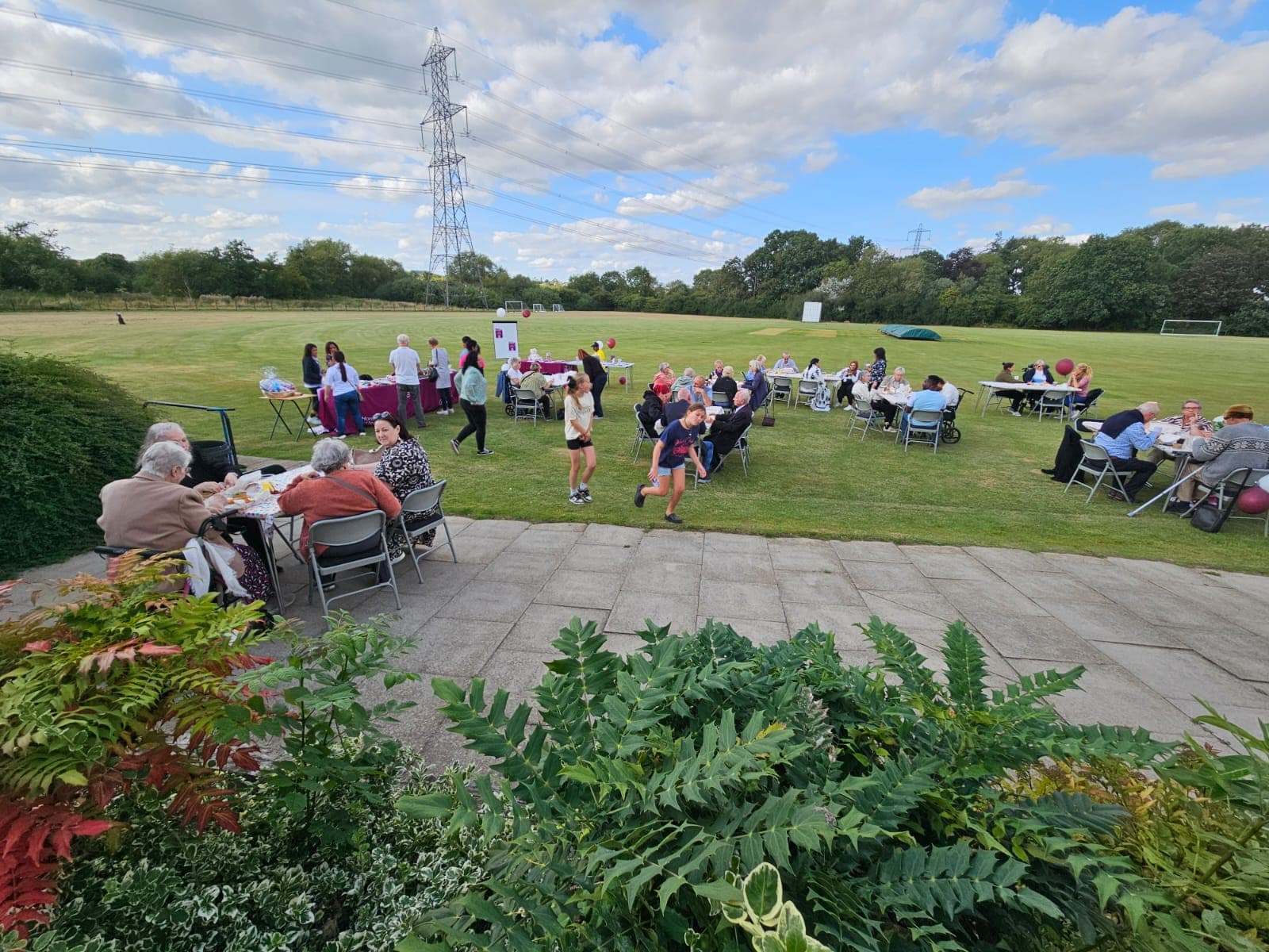 A community gathering on a grassy field with tables, chairs, and people enjoying food and conversation under a cloudy sky. - Home Instead