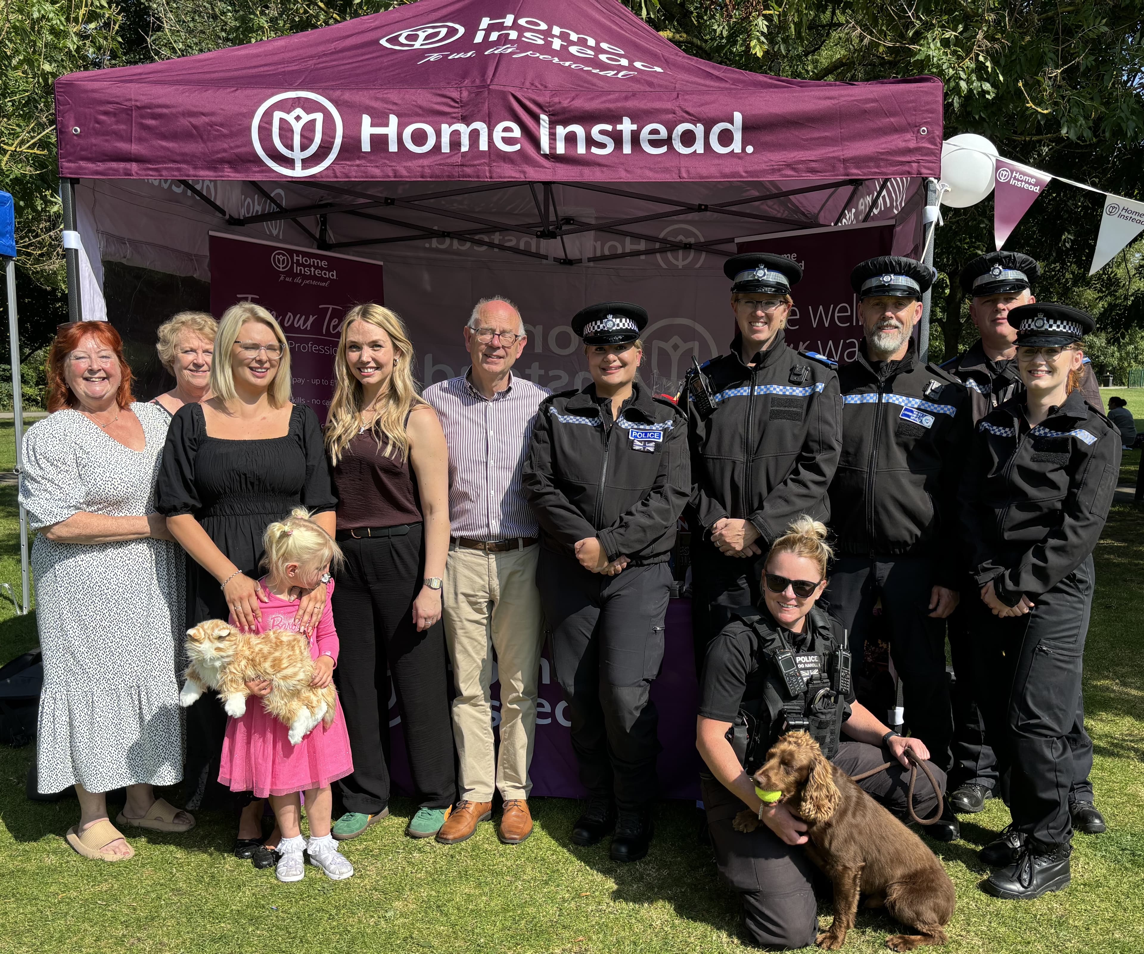 A group poses under a "Home Instead" canopy. Some are wearing police uniforms, and two hold dogs. - Home Instead